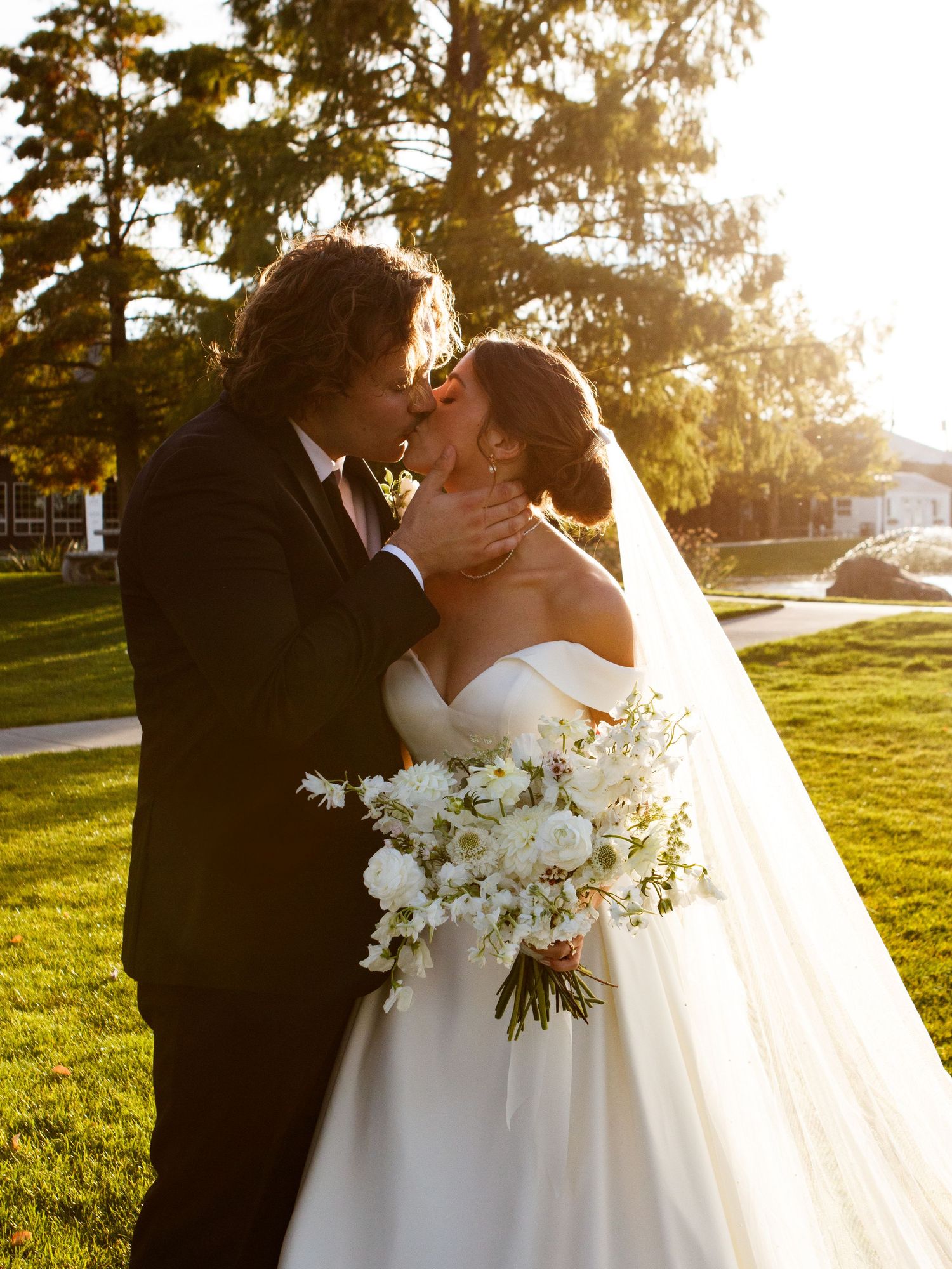 A couple shares a romantic kiss while holding a white floral bouquet during their sunset wedding ceremony on a grassy lawn.