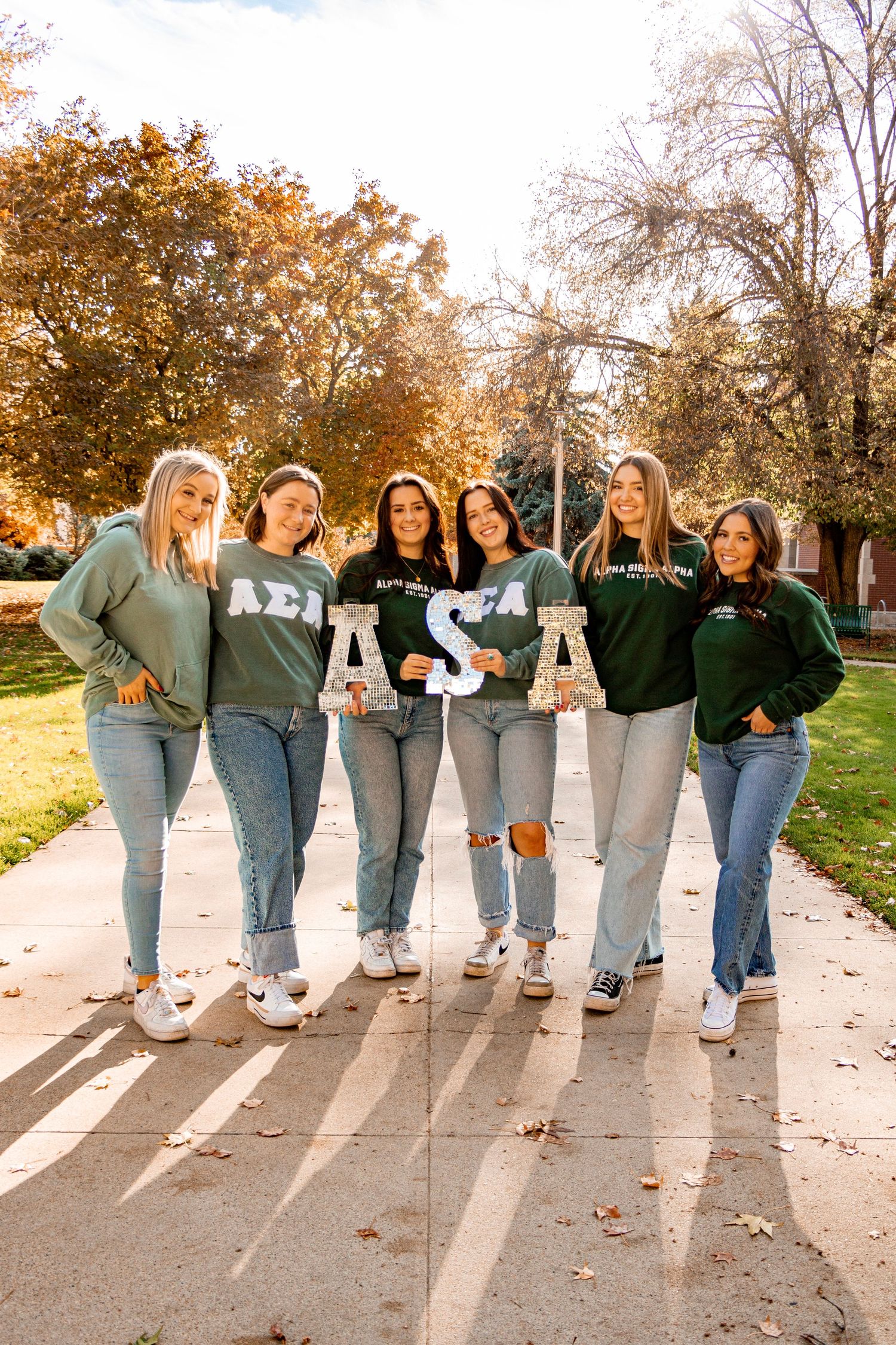 A group of students in green sweatshirts walk together on a campus path on a sunny autumn day.