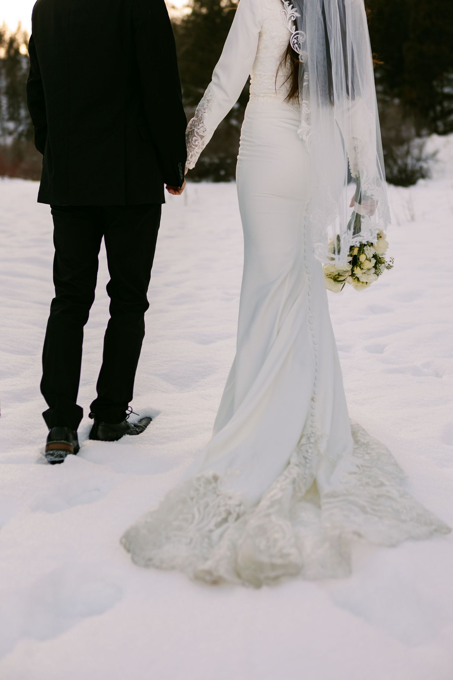A couple stands in snow during a winter wedding, with bride in white gown holding bouquet and groom in black suit.