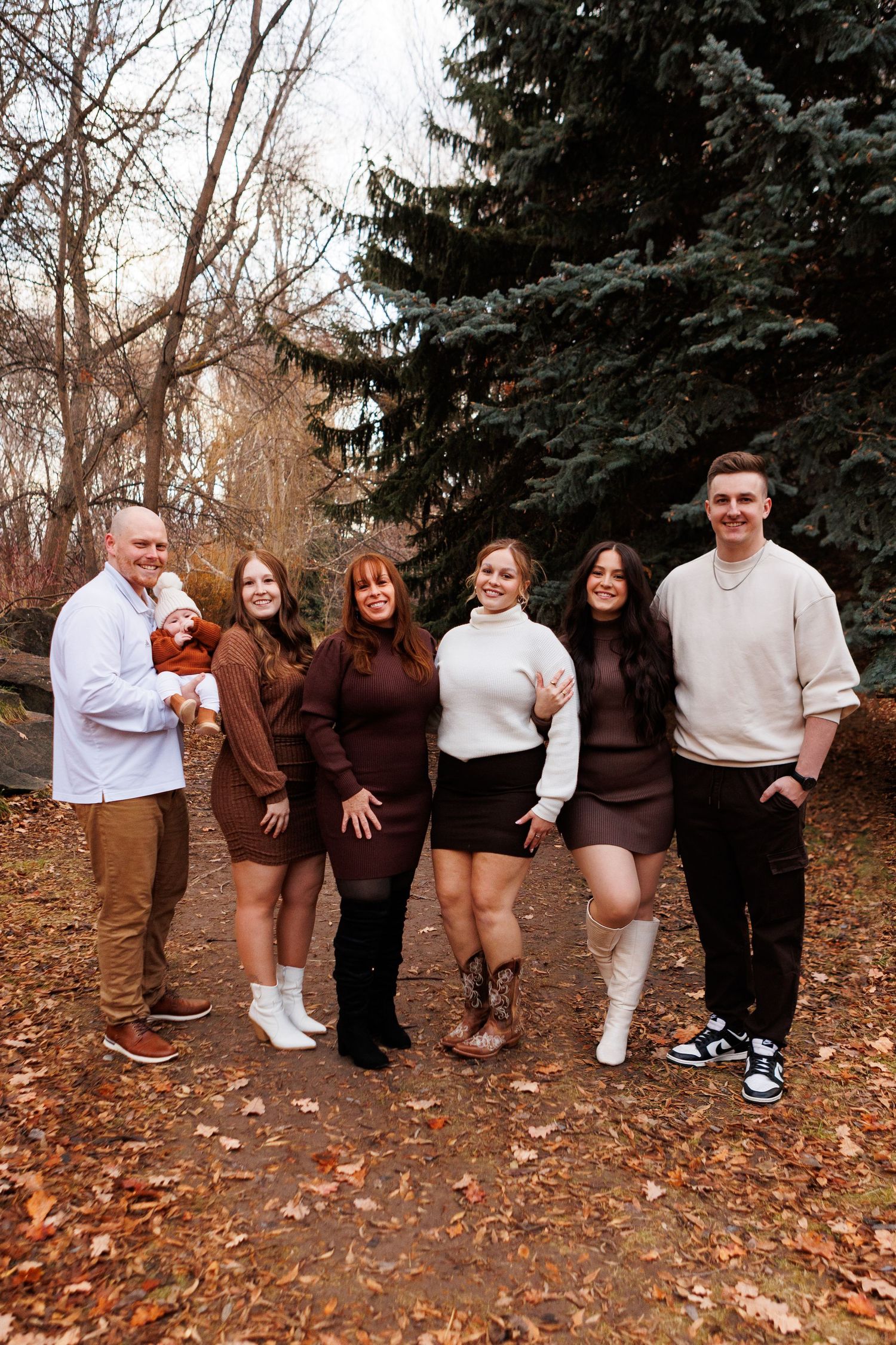 Group in coordinated brown and white outfits pose together on fallen autumn leaves with evergreen trees in background.