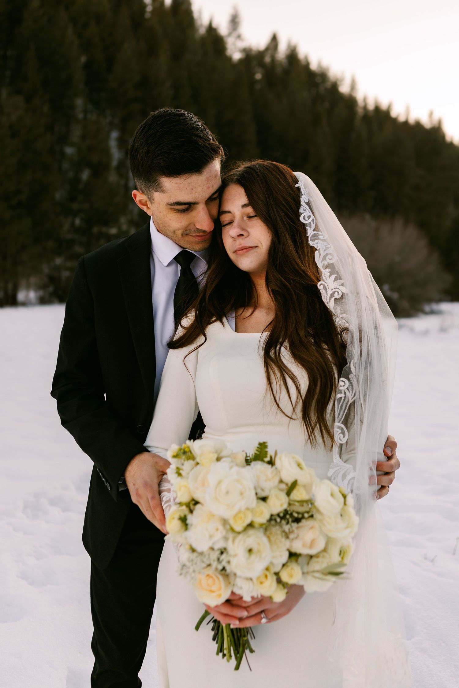 A romantic winter wedding photo featuring a couple embracing in the snow with a white floral bouquet and forest backdrop.