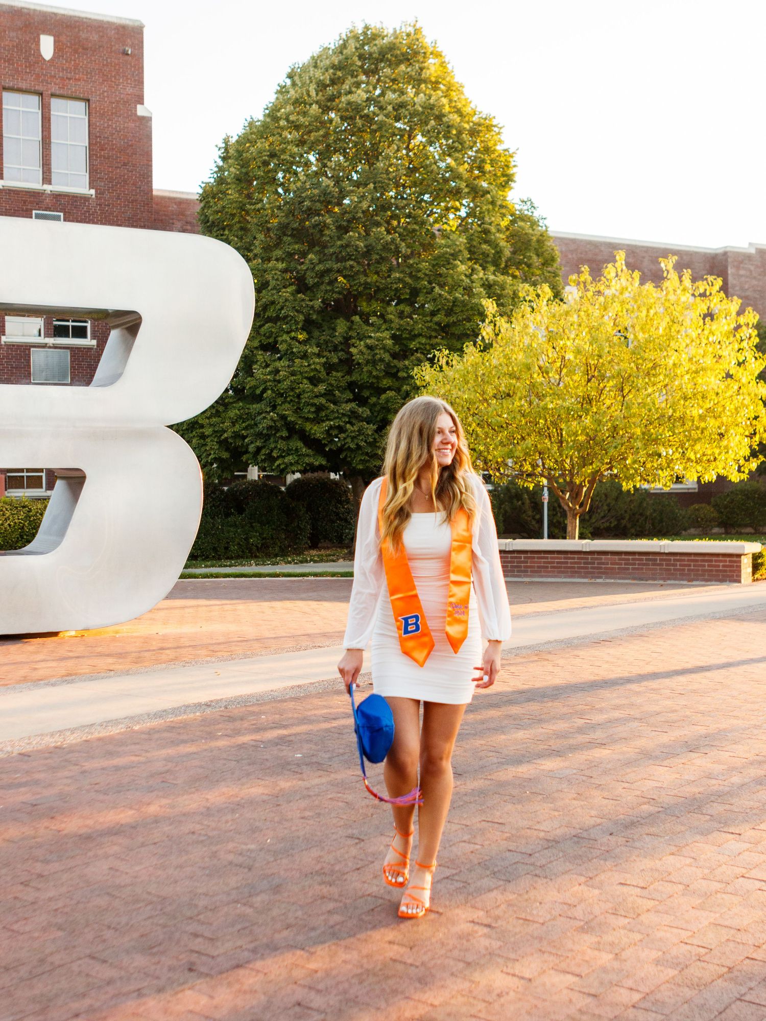 A figure in a white dress and orange vest walks outdoors on a sunny day near autumn trees and a large letter B sculpture.