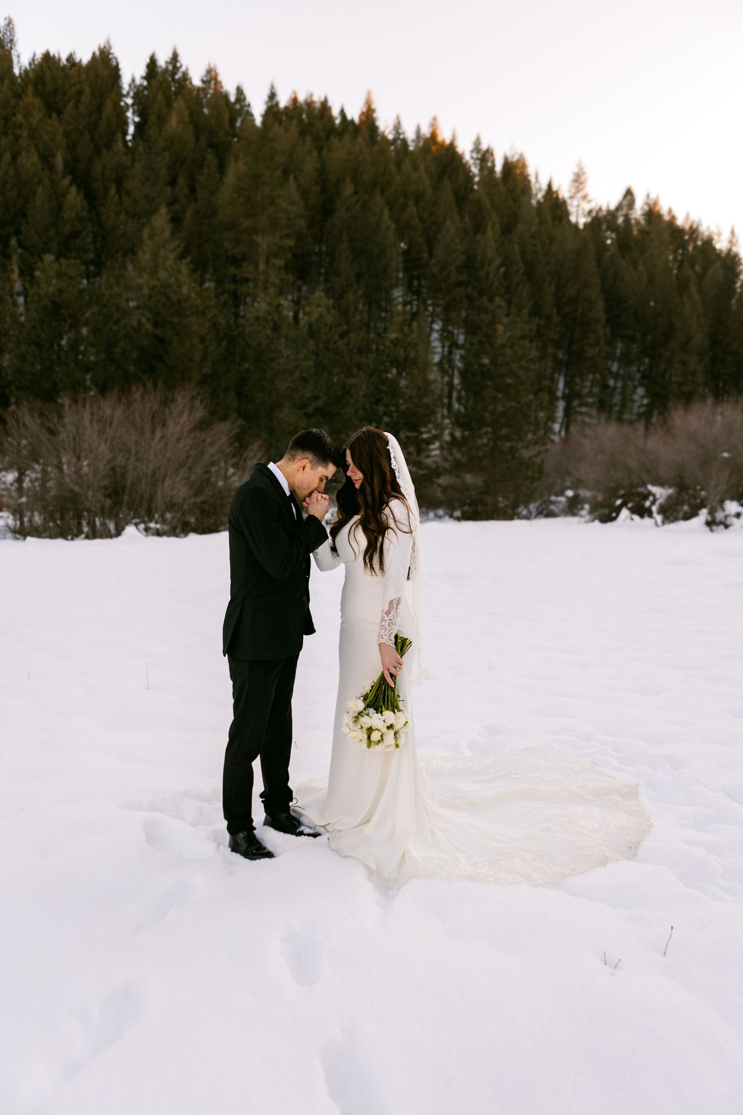 Two people stand together in formal wedding attire against a snowy forest backdrop on their winter wedding day.