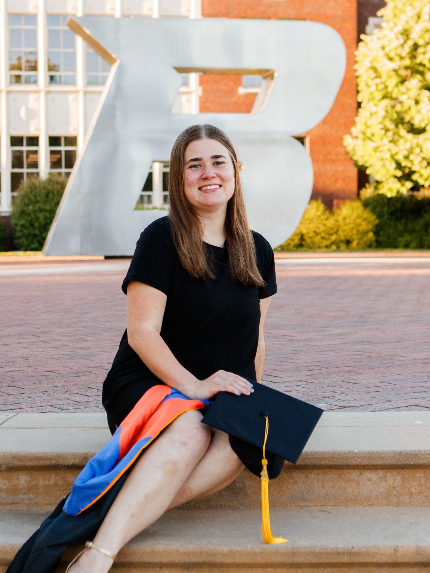 Graduate sits on stone steps near a large decorative B letter sculpture wearing cap and gown with black dress.