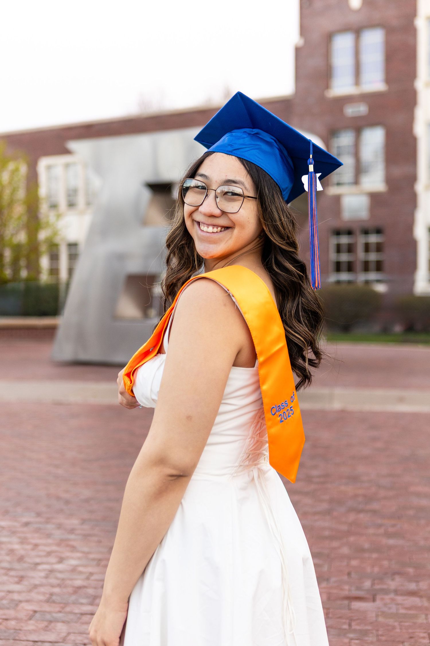 Graduate in white dress and blue cap with orange honors stole poses on campus brick courtyard at academic building.