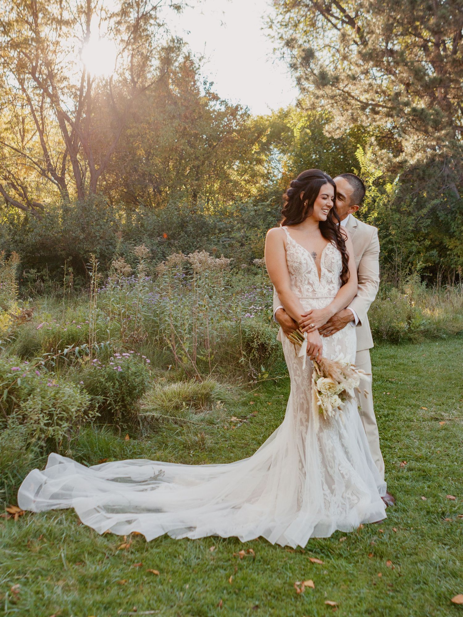 Two people in wedding attire embrace in a sun-dappled outdoor garden setting with a flowing white dress train.