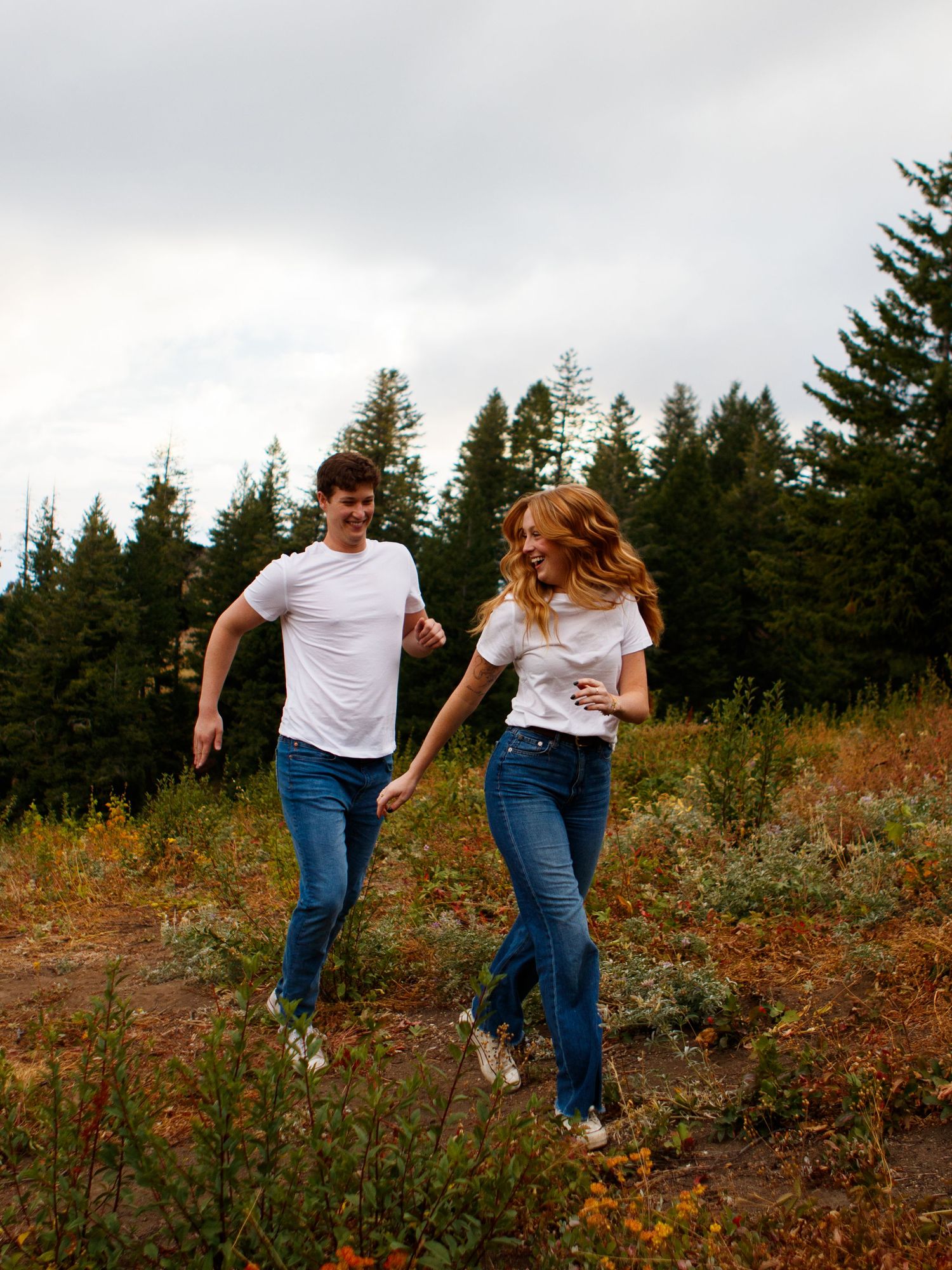 Two people in matching white t-shirts and jeans running joyfully through a hillside meadow with evergreen trees in background.