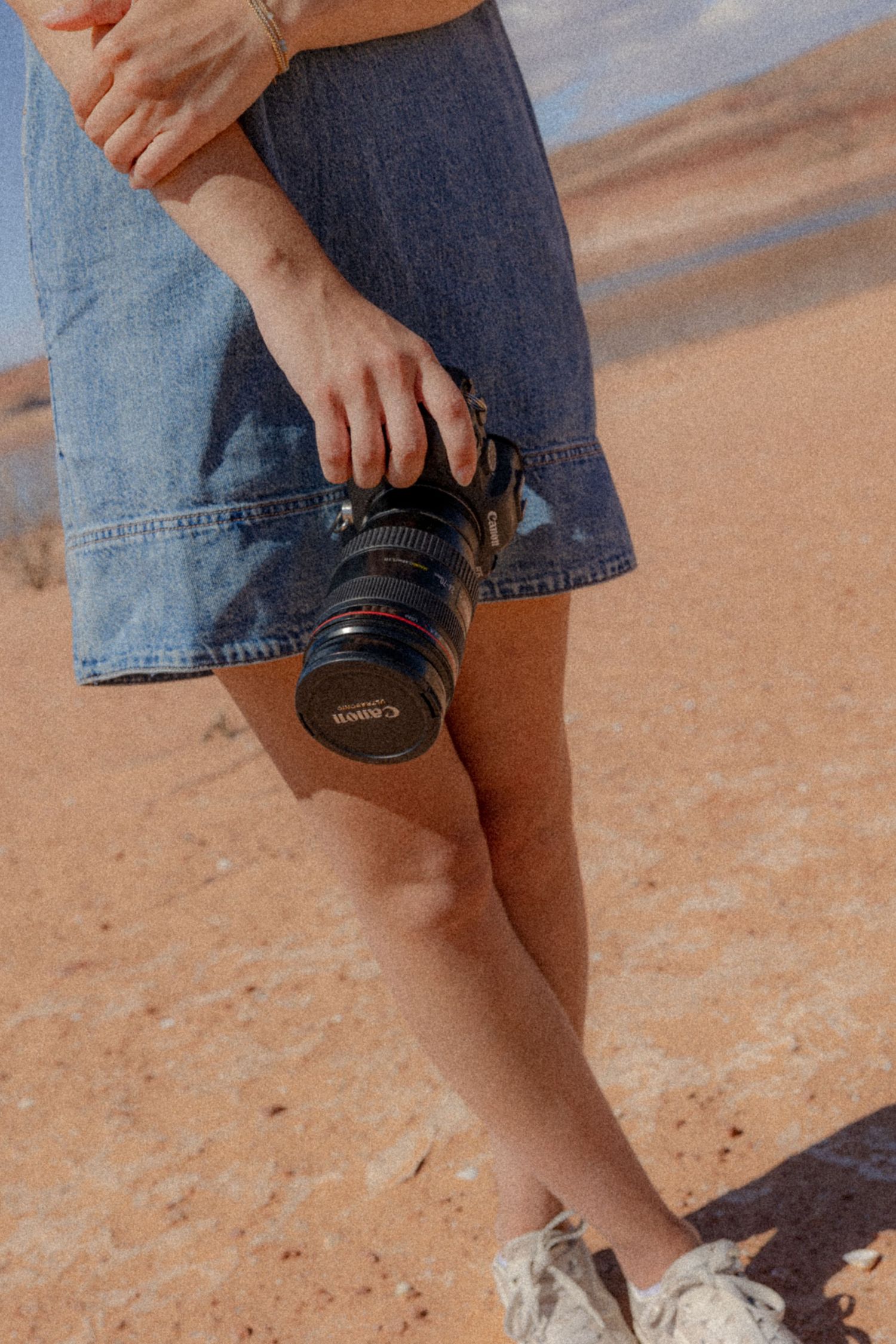 Person in blue jeans holds a professional camera with lens while standing outdoors on reddish ground.