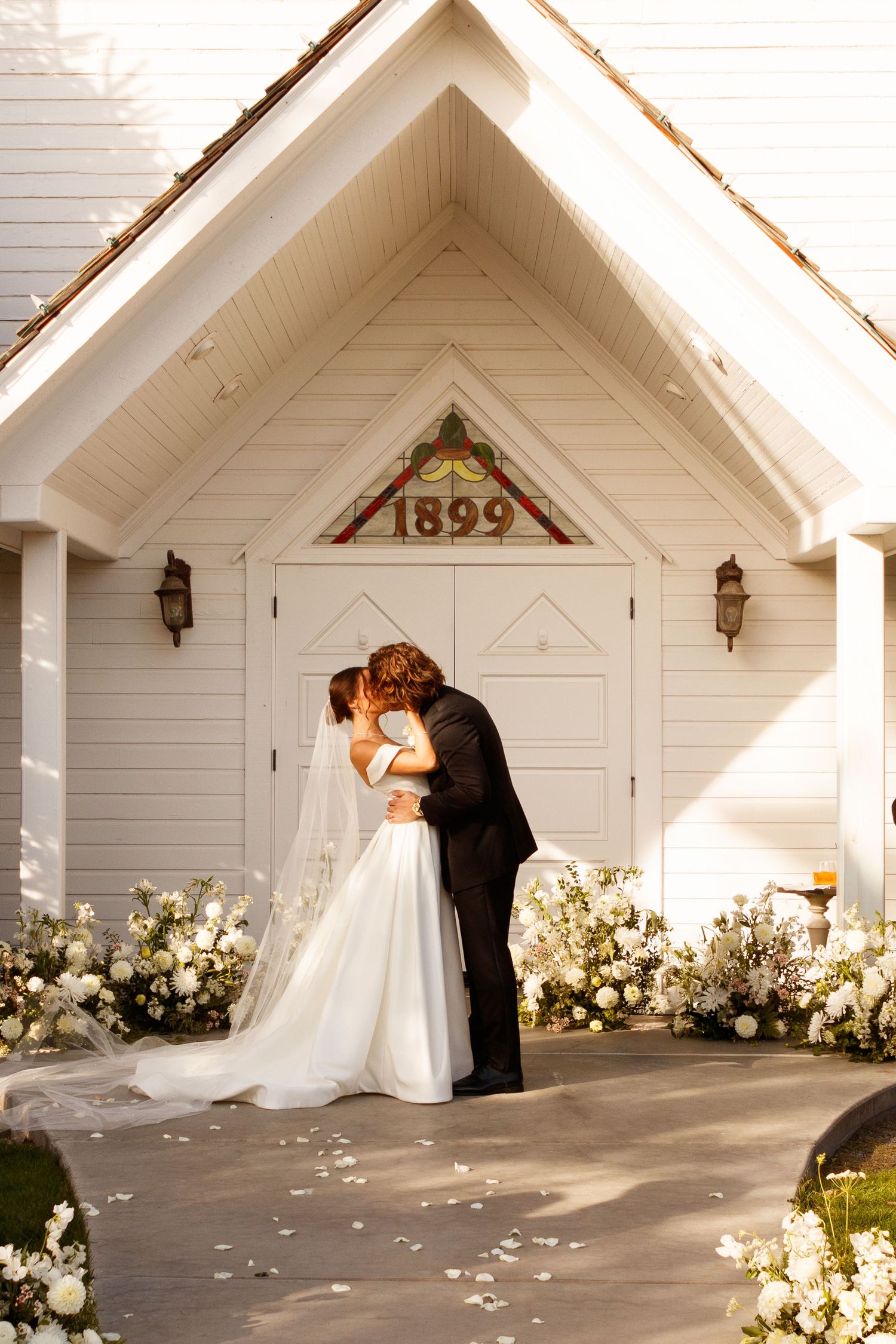 A romantic wedding couple embraces in front of a white chapel entrance adorned with floral arrangements and the year 1899.