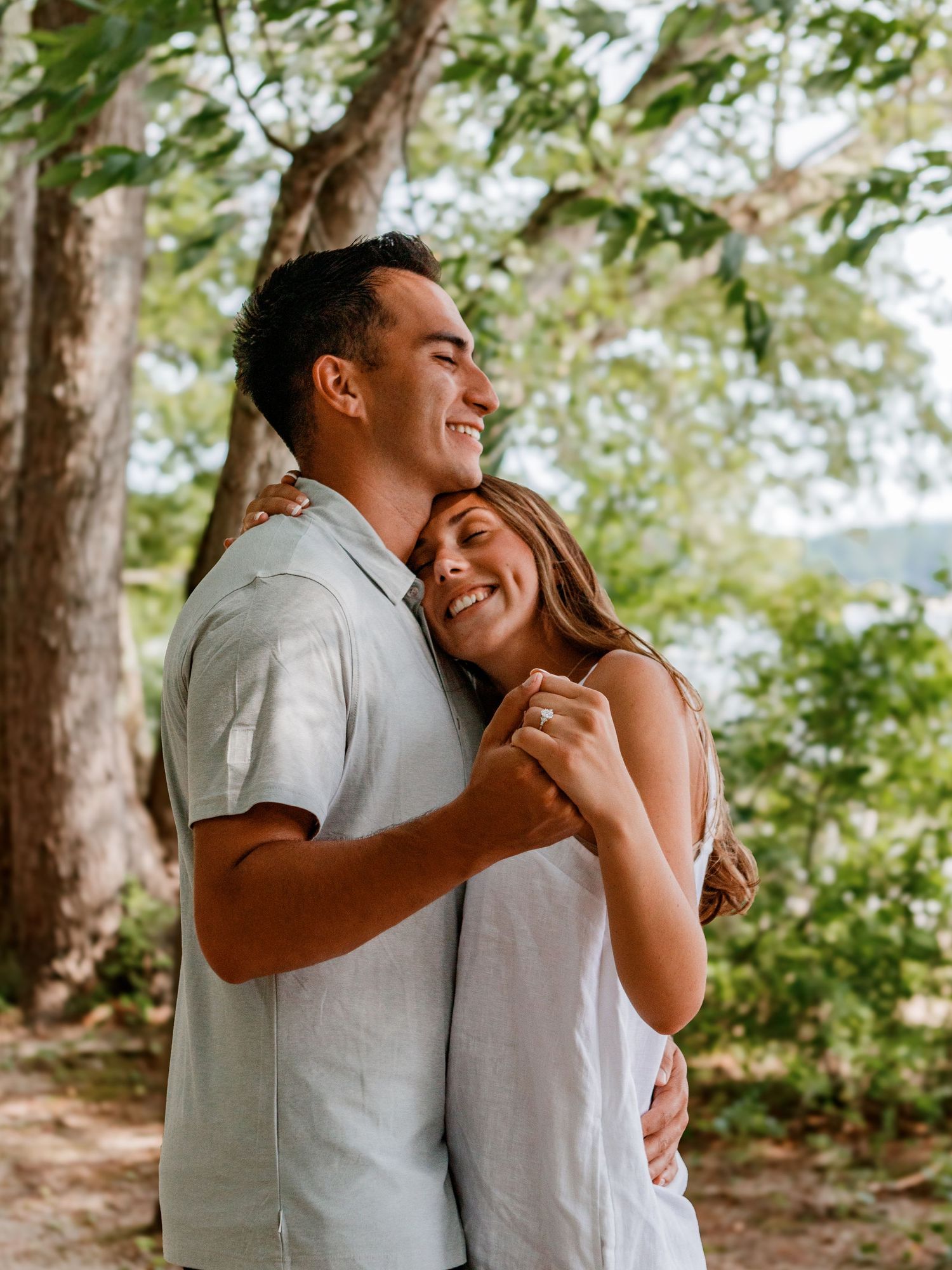 Two people share a tender romantic embrace while standing outdoors among trees on a sunny day.