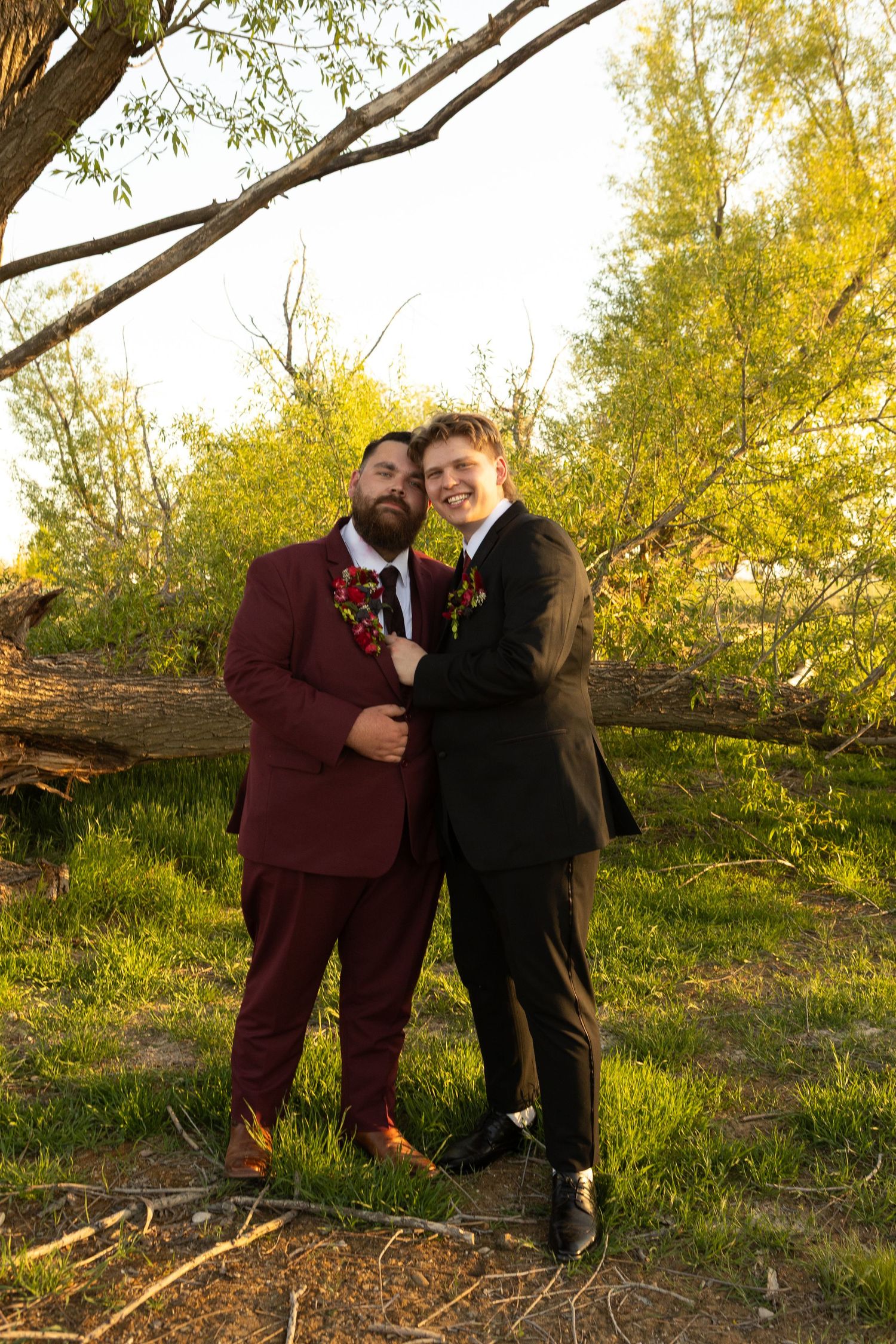 A couple in burgundy and black formal suits embracing outdoors surrounded by autumn trees in golden light.