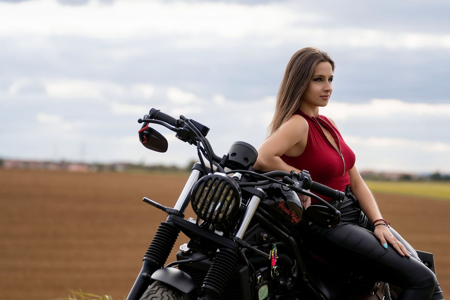 Young woman in red top leaning on black motorcycle in open field.
