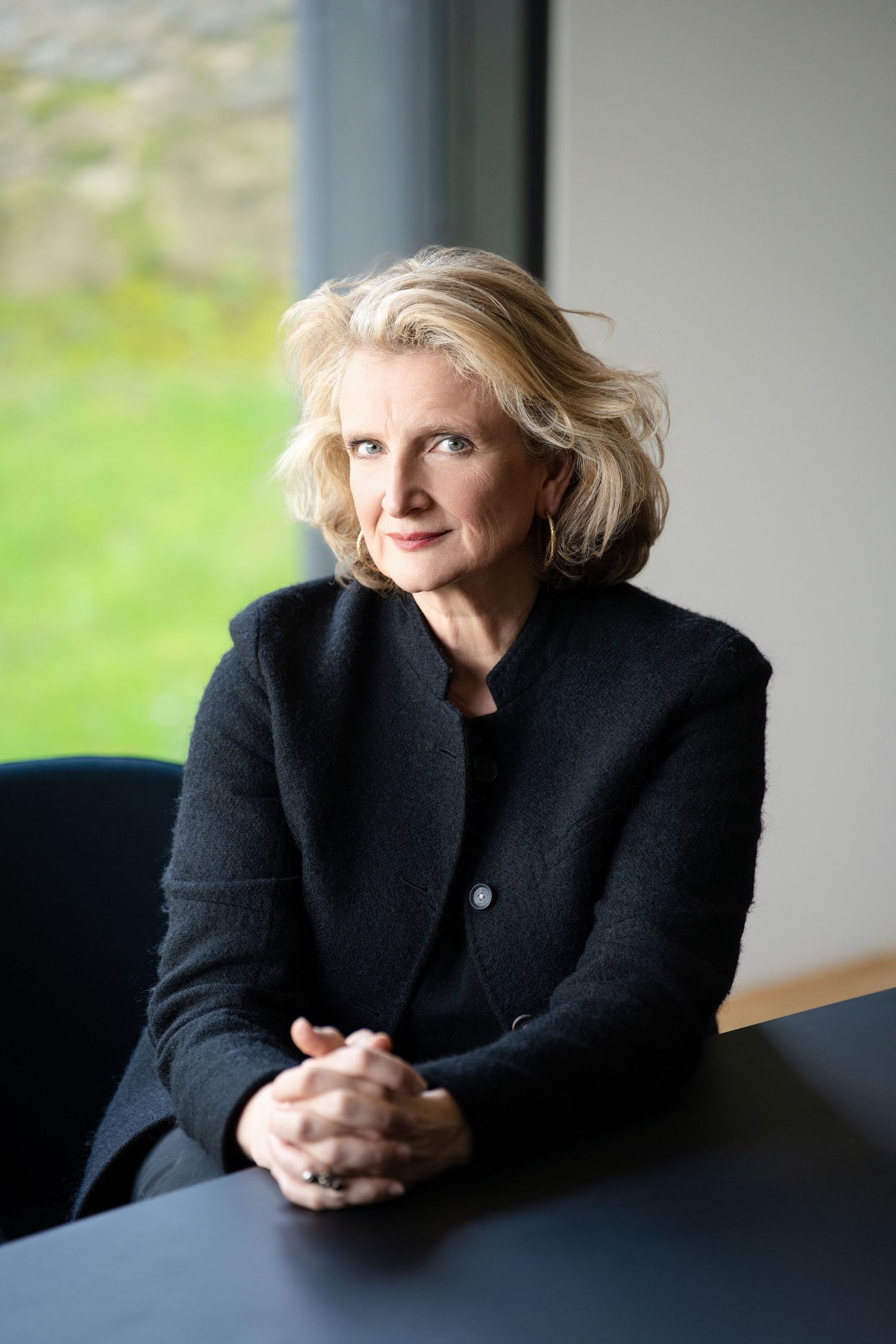Elegant blonde woman in black jacket sitting at desk, gazing thoughtfully toward window.