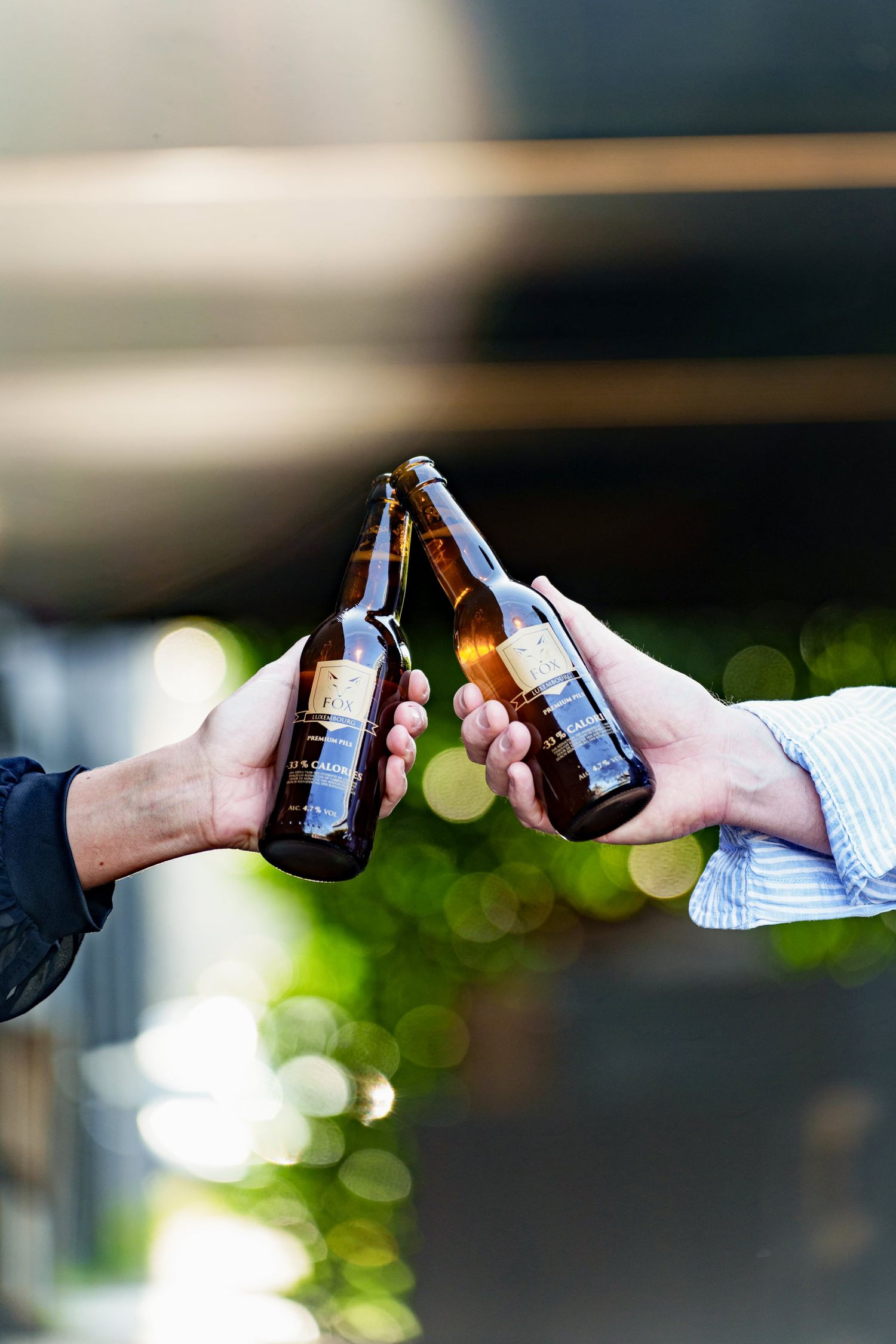 Two people clinking brown beer bottles together outdoors with a bokeh green background.