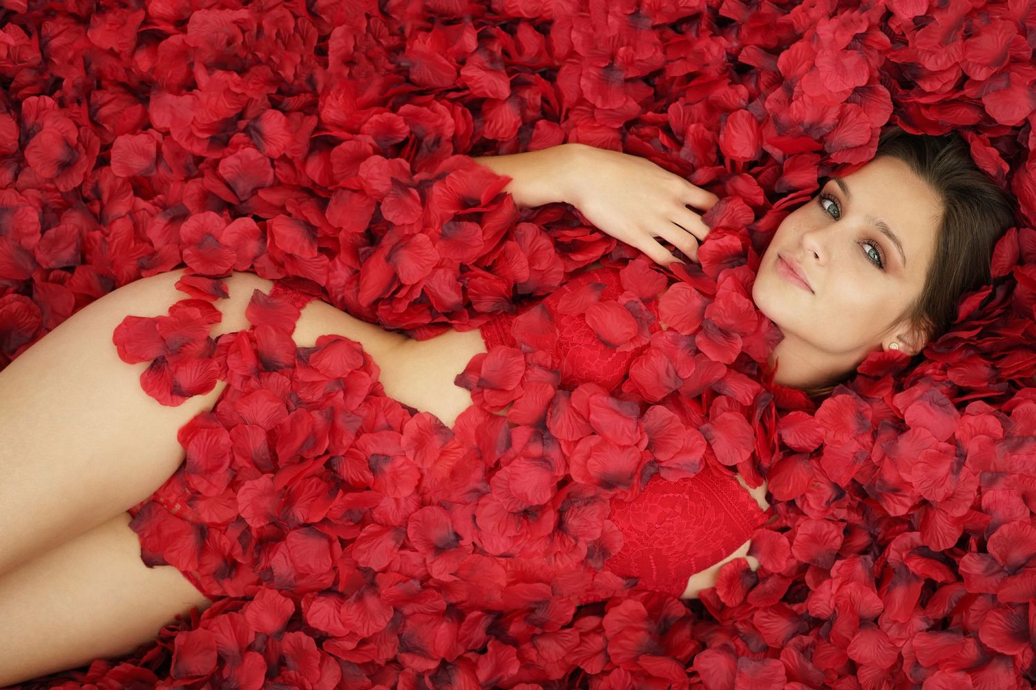 Young woman lying in a bed of red rose petals in an artistic and romantic photography composition.