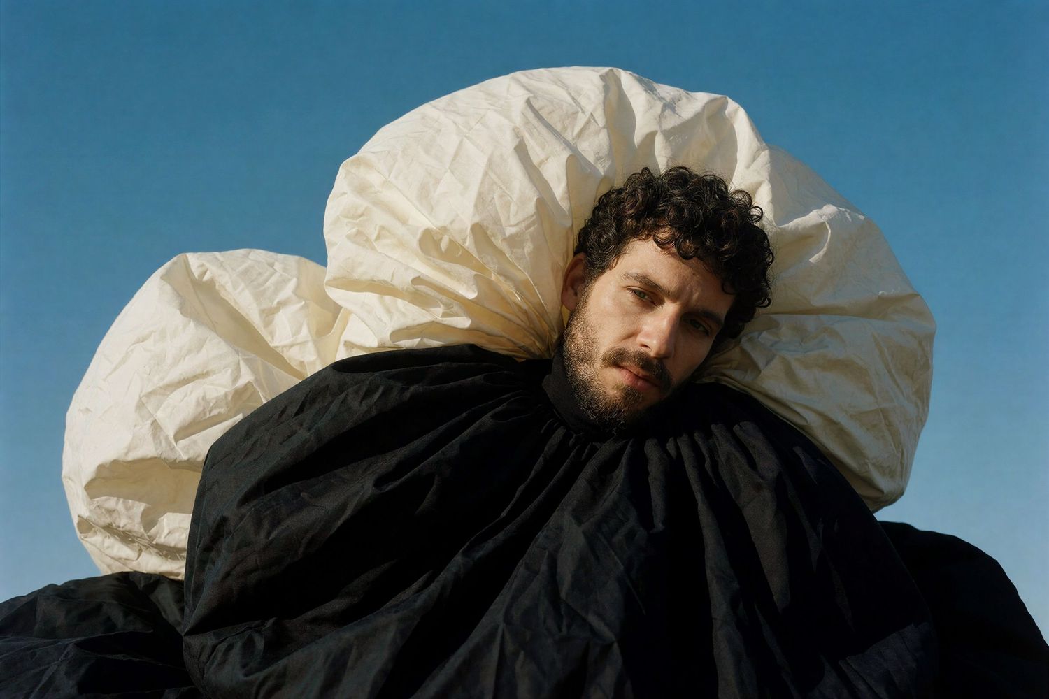 A bearded man wrapped in a large black and white inflatable sculptural garment outdoors.