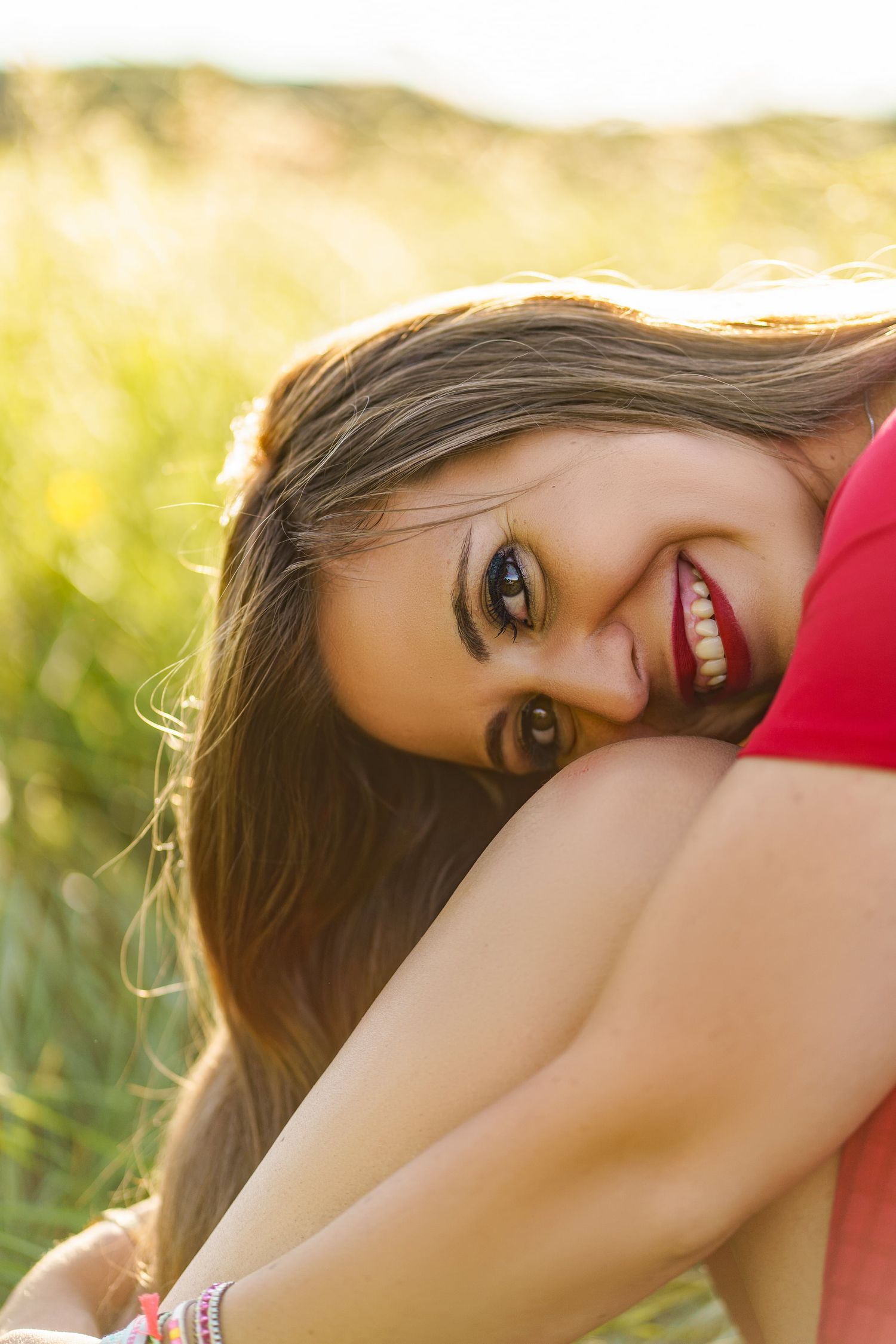 Happy young woman with long brown hair smiling while sitting in a sunny green meadow wearing red.
