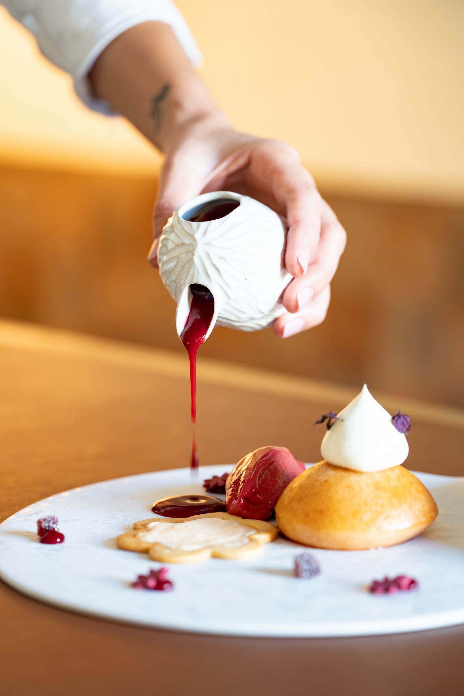 Chef pouring red berry sauce from decorative white pitcher onto a gourmet dessert plate.