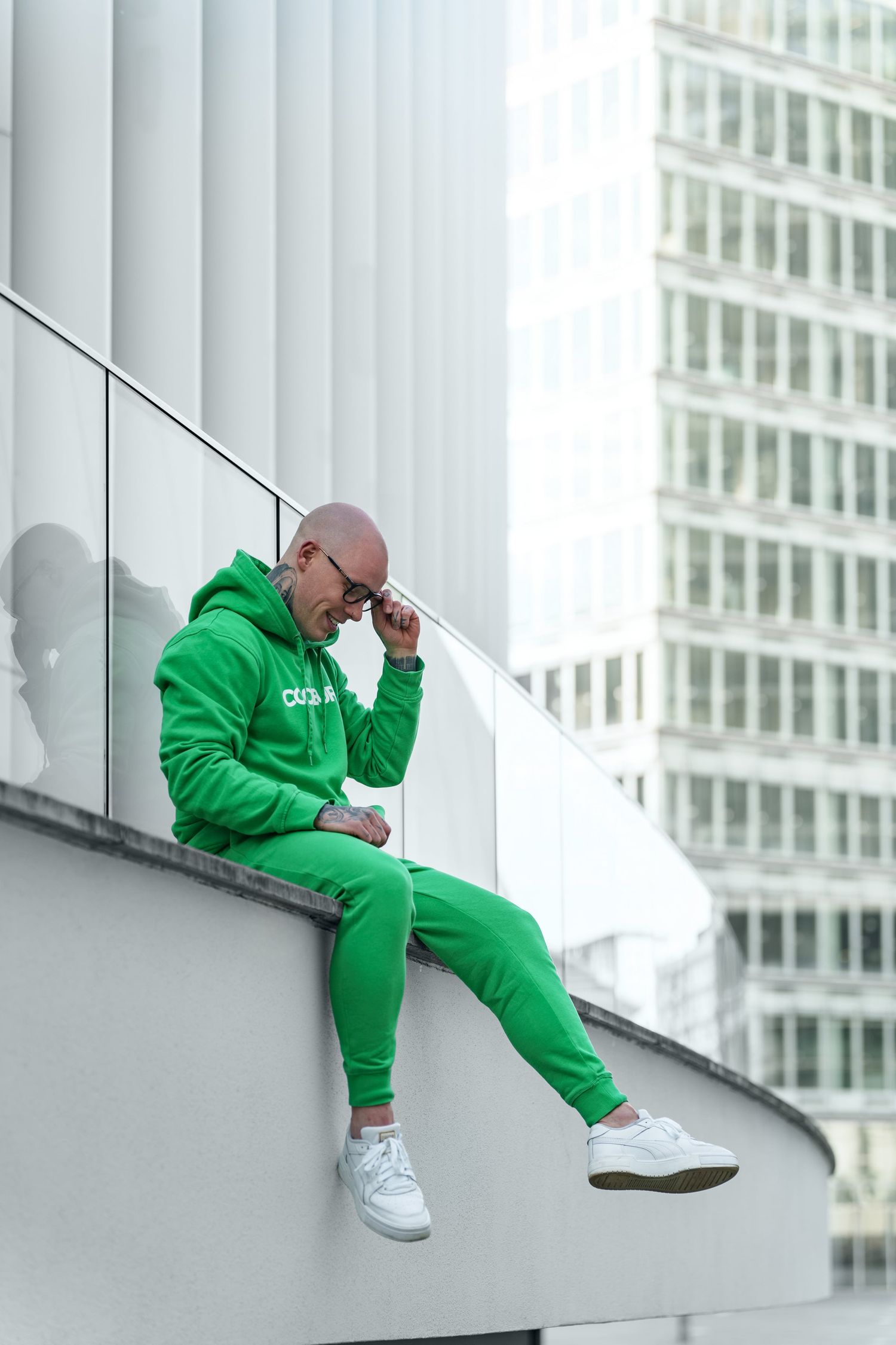 Bald man in bright green hoodie and joggers sitting on urban ledge with city buildings in background.