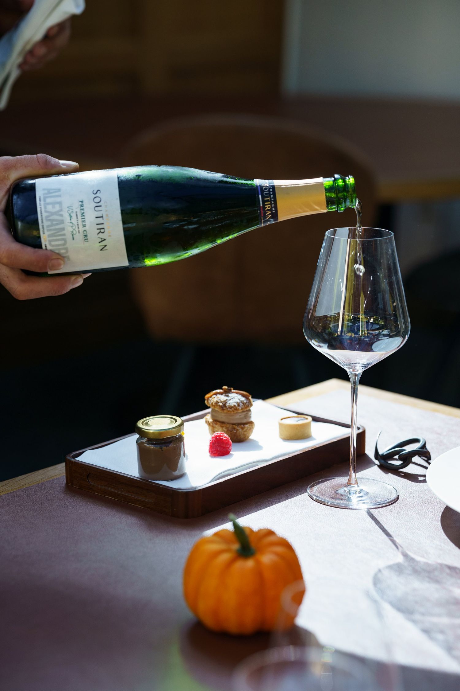 Hand pouring sparkling wine into elegant glass beside autumn dessert board with small pumpkin decoration.