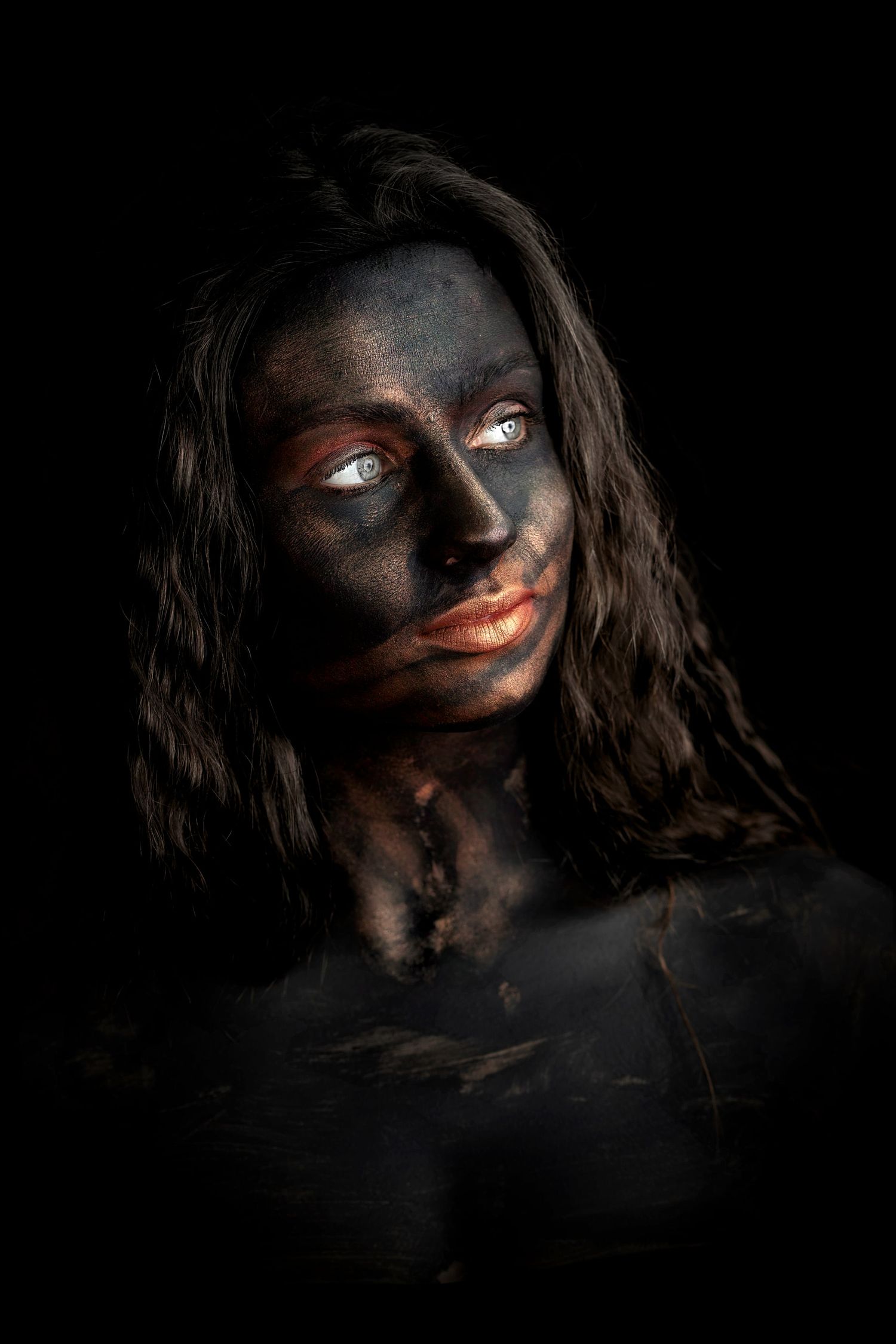 Woman with dark body paint and striking blue eyes posing against a black background.