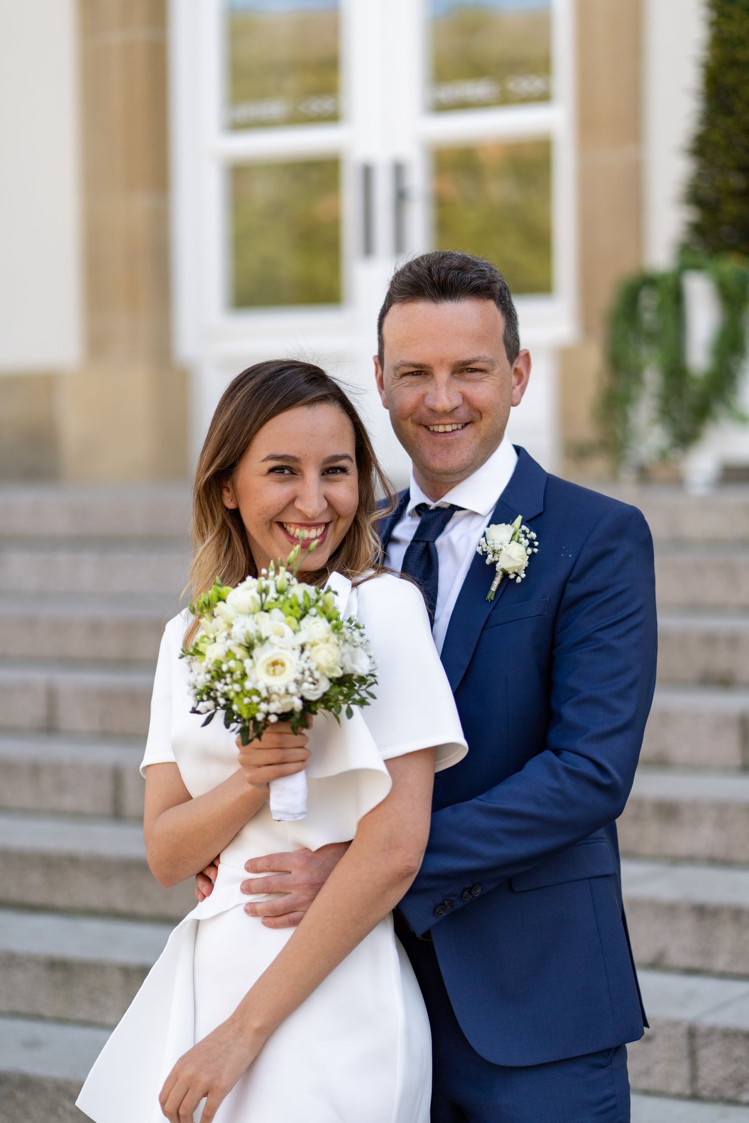 Happy bride in white dress holding bouquet embraced by groom in navy suit on church steps.