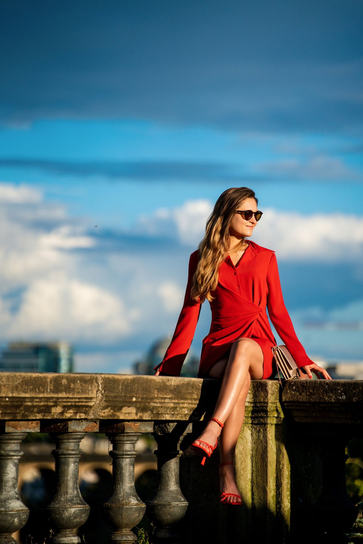 An elegant woman in a red wrap dress and sunglasses sits on a stone balustrade against a dramatic blue sky.