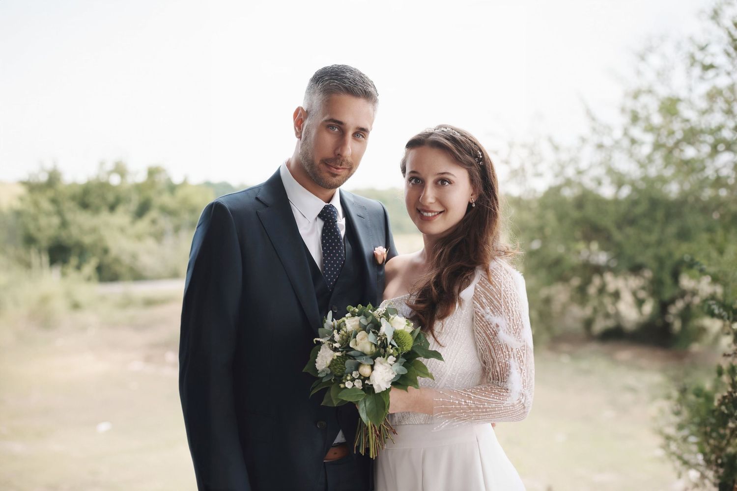 Smiling bride and groom pose together outdoors holding a white floral bouquet on their wedding day.