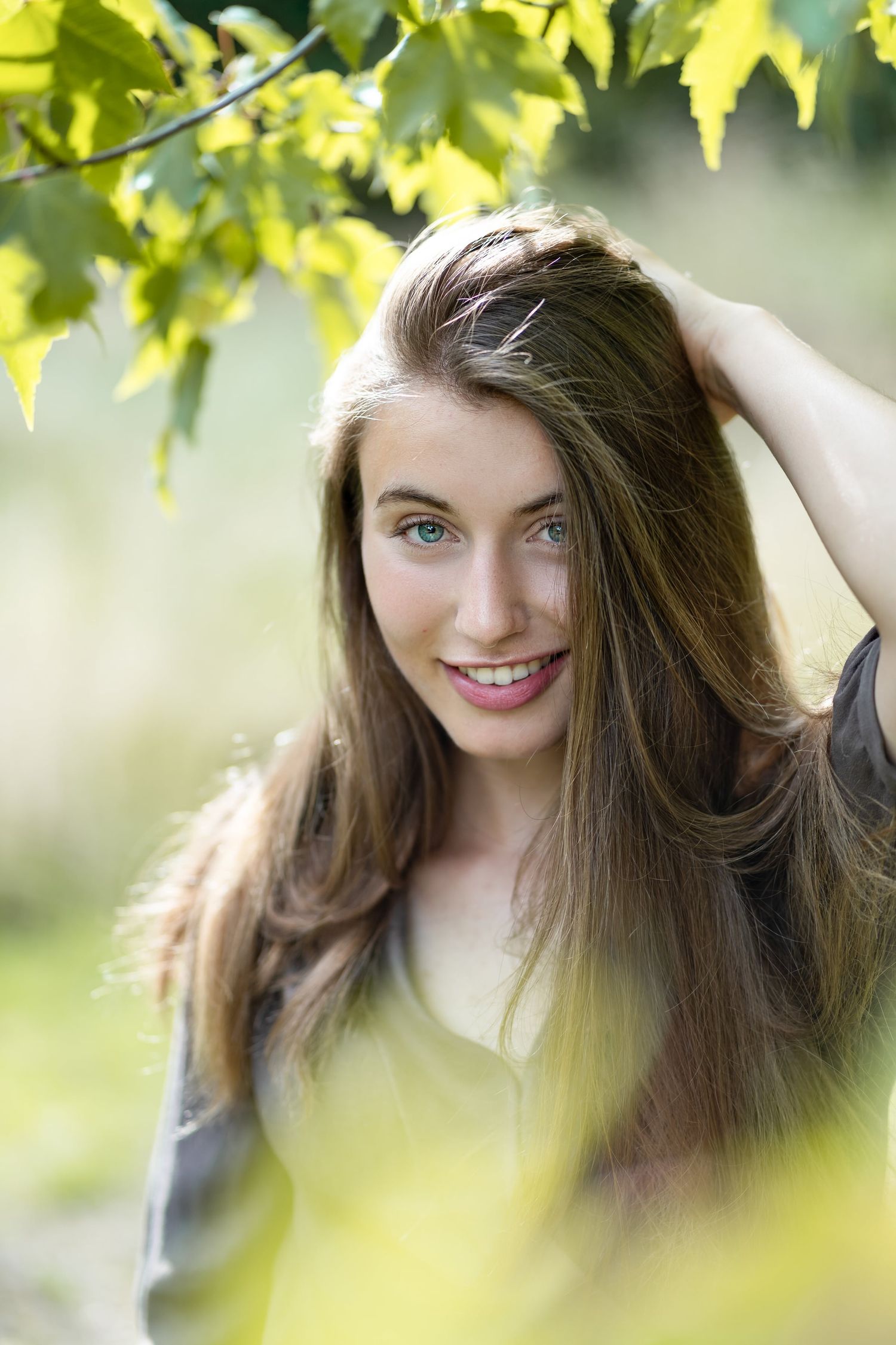 Young woman with long brown hair and blue eyes smiling under green maple leaves in a park.