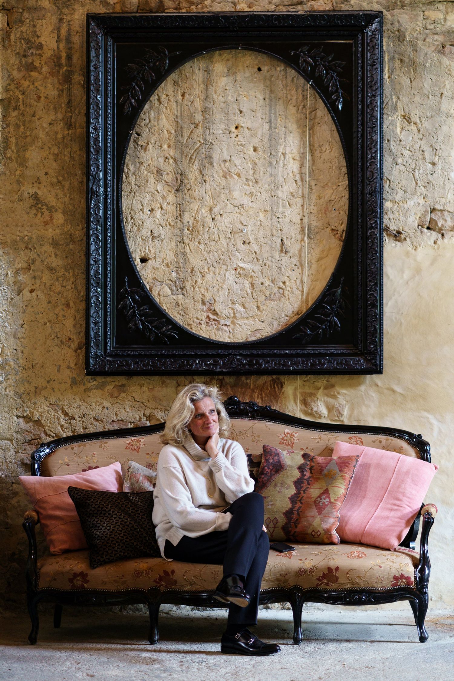 Mature blonde woman sitting on ornate vintage sofa beneath large decorative frame.