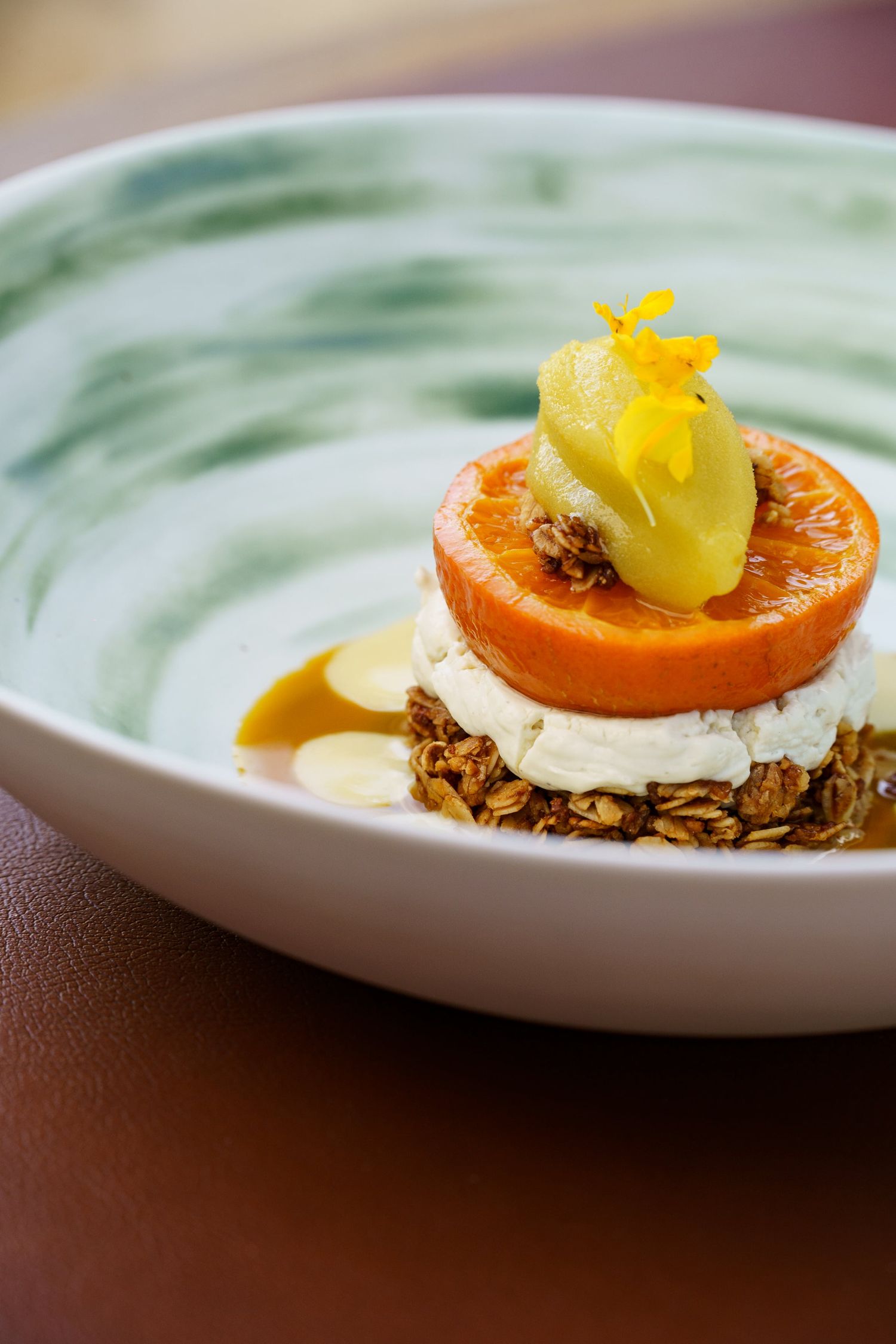 A refined dessert with granola base, cream, caramelized orange slice, and yellow flower petal garnish in a bowl.