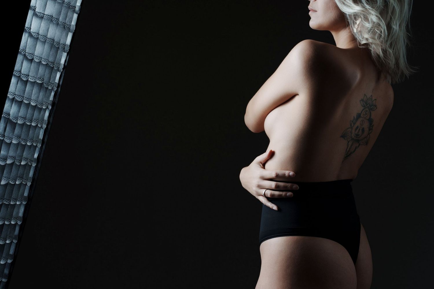Side profile of a tattooed blonde woman posing topless in black underwear against a dark studio background.