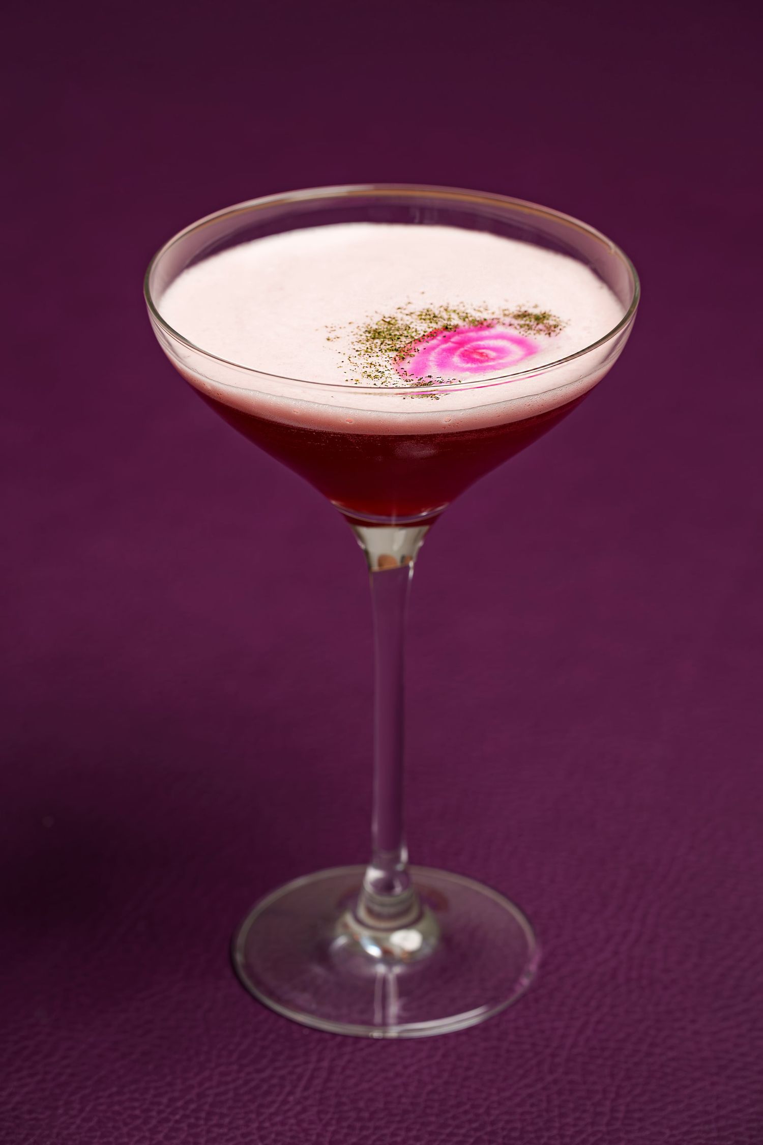 An elegant pink cocktail in a martini glass garnished with a rose petal on a purple background.