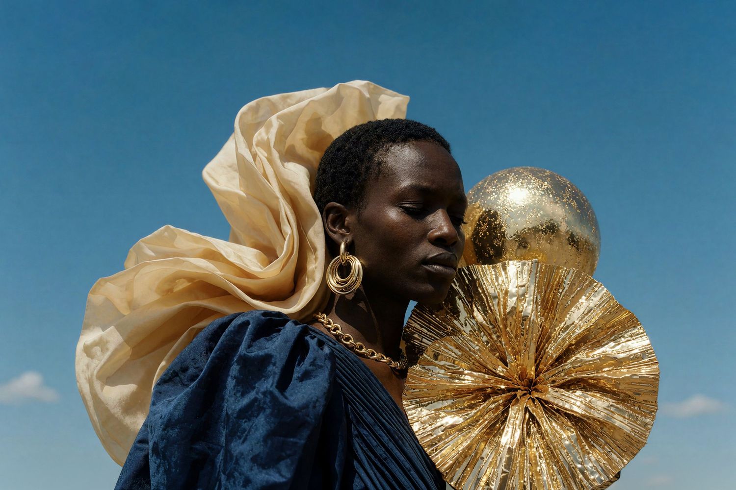 Fashion portrait of a woman in blue dress with gold accessories and oversized fan against blue sky.