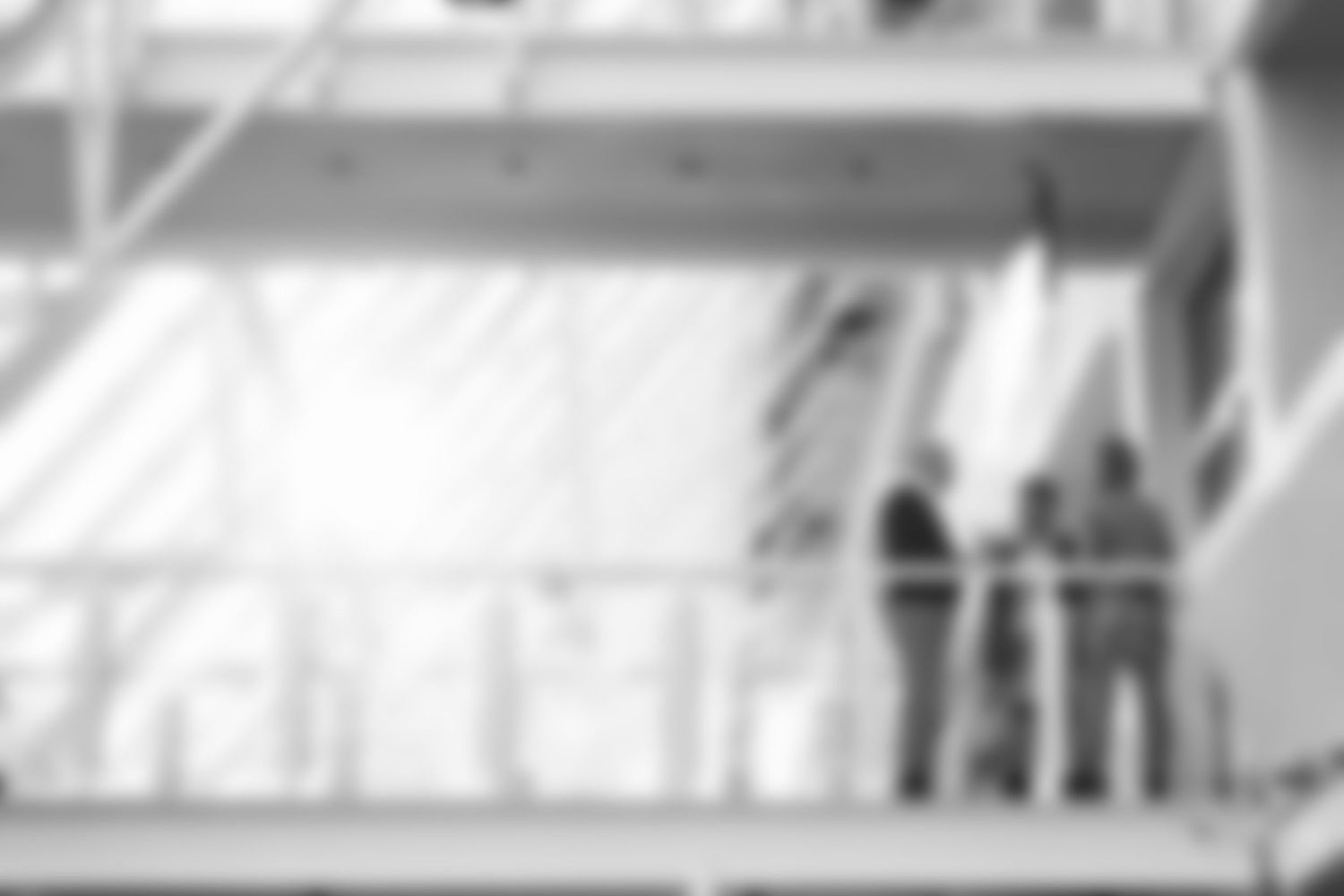 Blurred black and white image of three people talking on a bright modern building balcony.