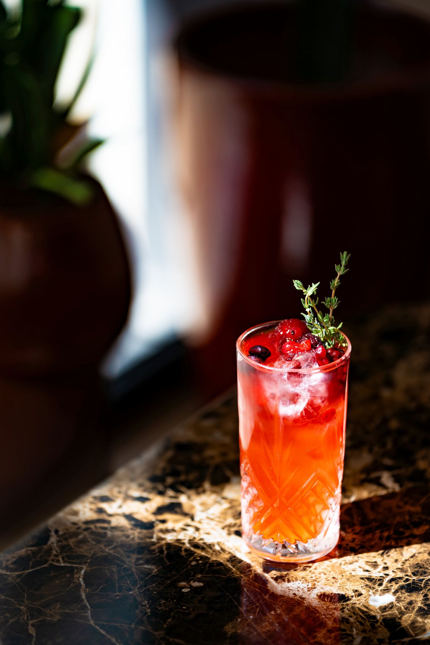 Vibrant red berry cocktail with thyme garnish in crystal highball glass on marble bar.