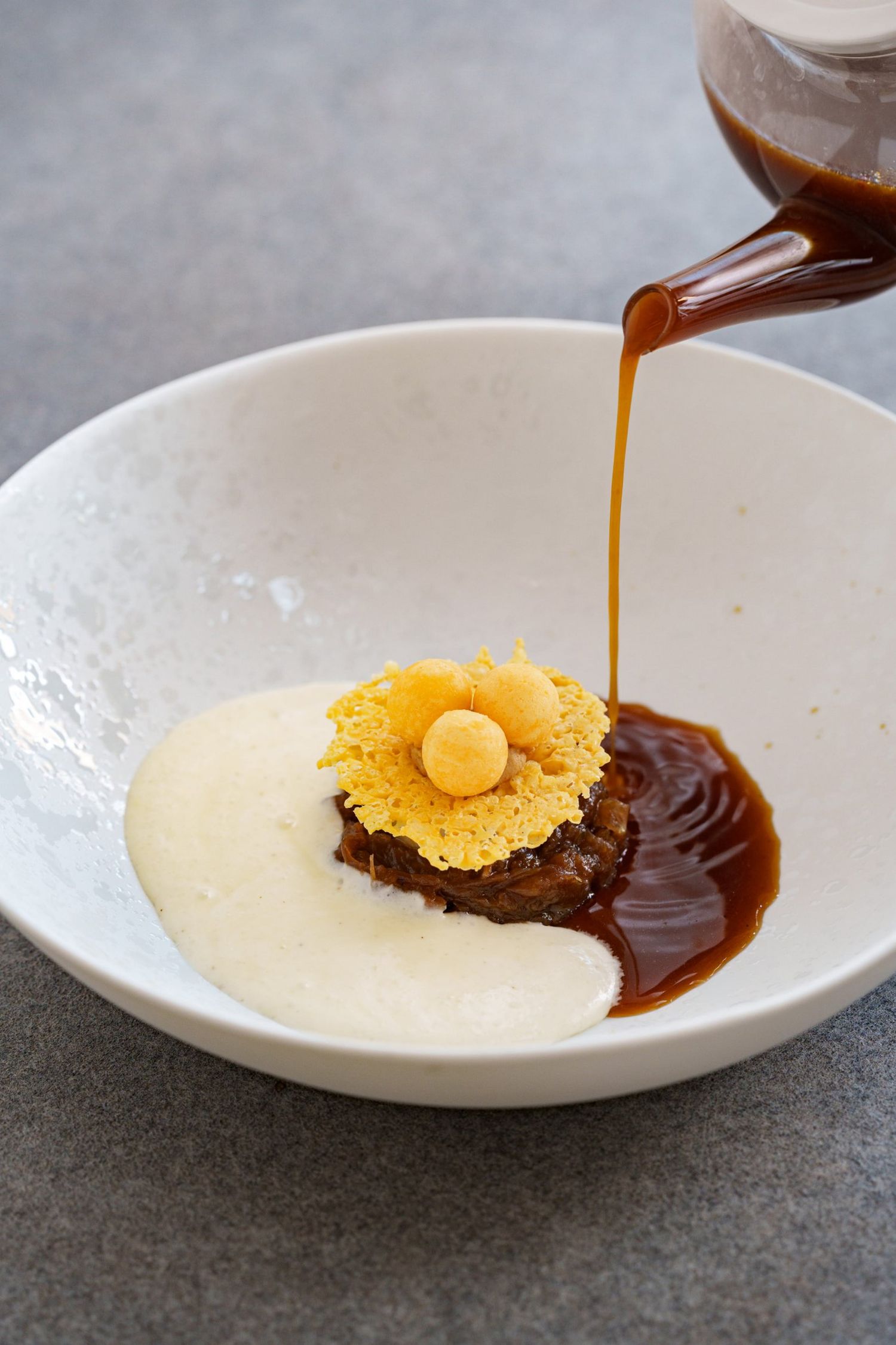 A gourmet plated dish with rich brown sauce being poured over a delicate nest garnish in a white bowl.