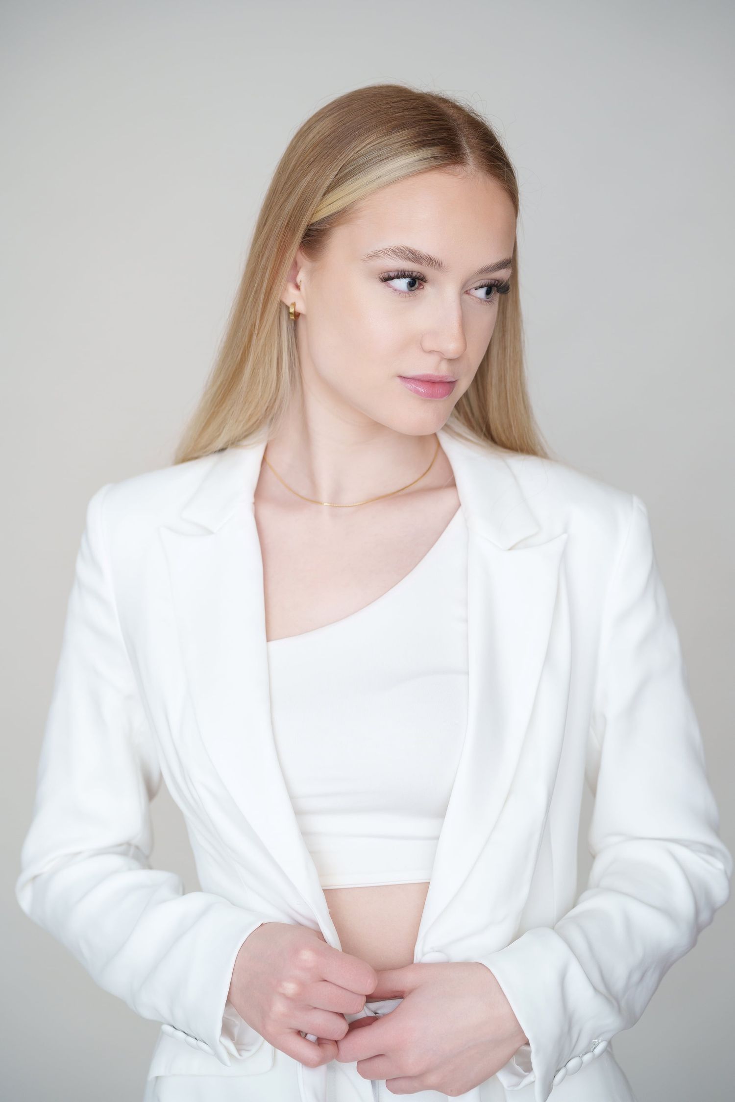 Young blonde woman in white blazer and crop top posing in clean studio setting.