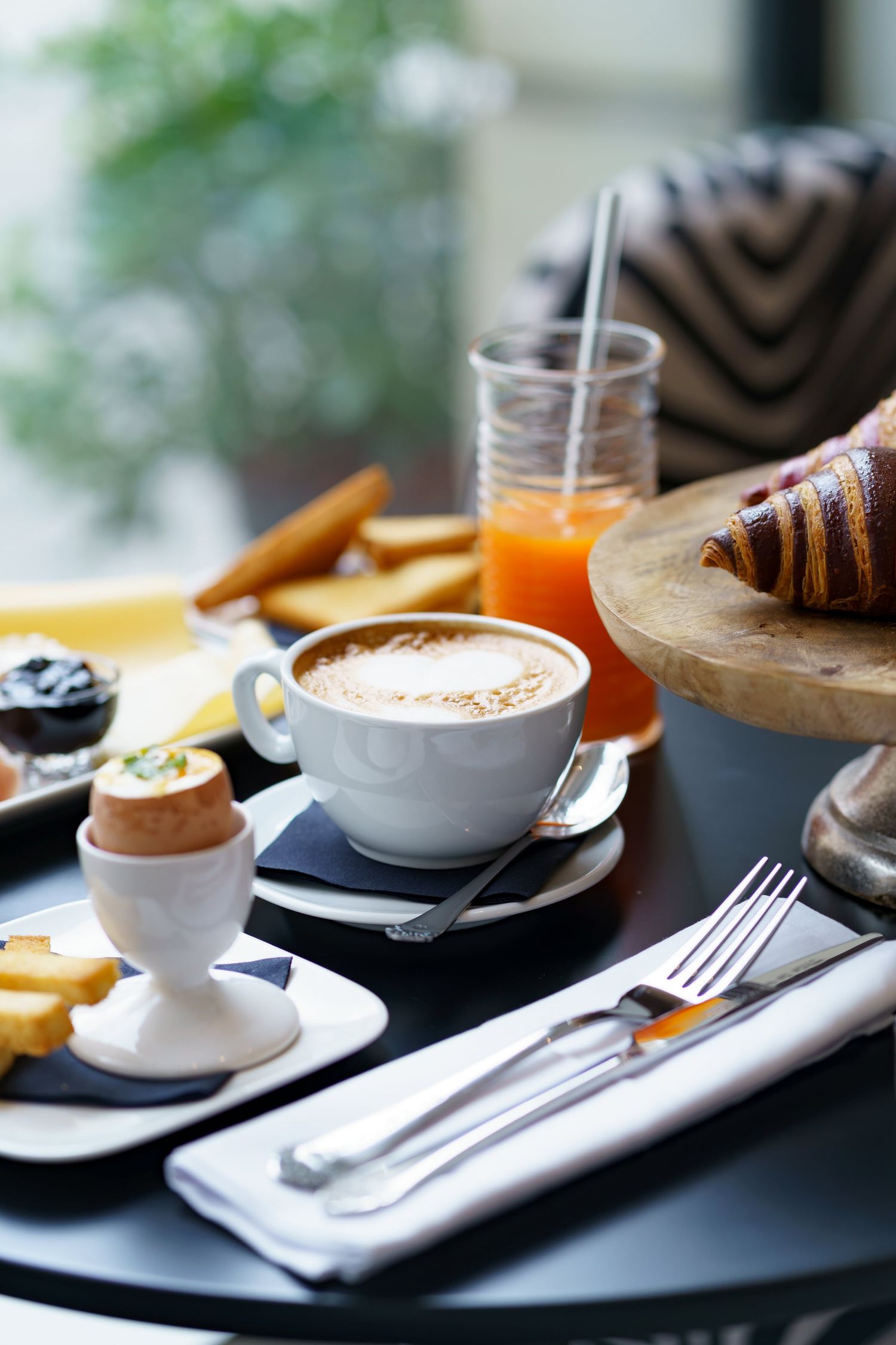 A classic French breakfast spread with cappuccino, soft boiled egg, croissant, orange juice, and jam on a dark table.