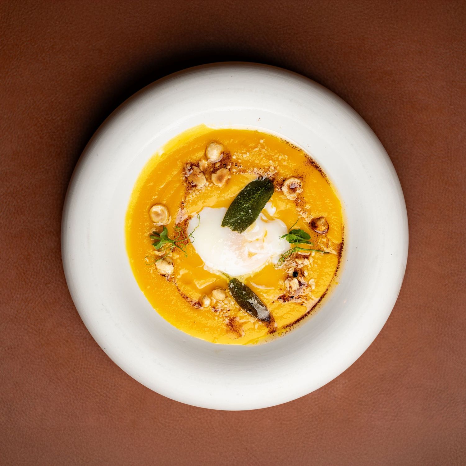 Overhead view of creamy pumpkin soup with poached egg, hazelnuts, and sage in white bowl.