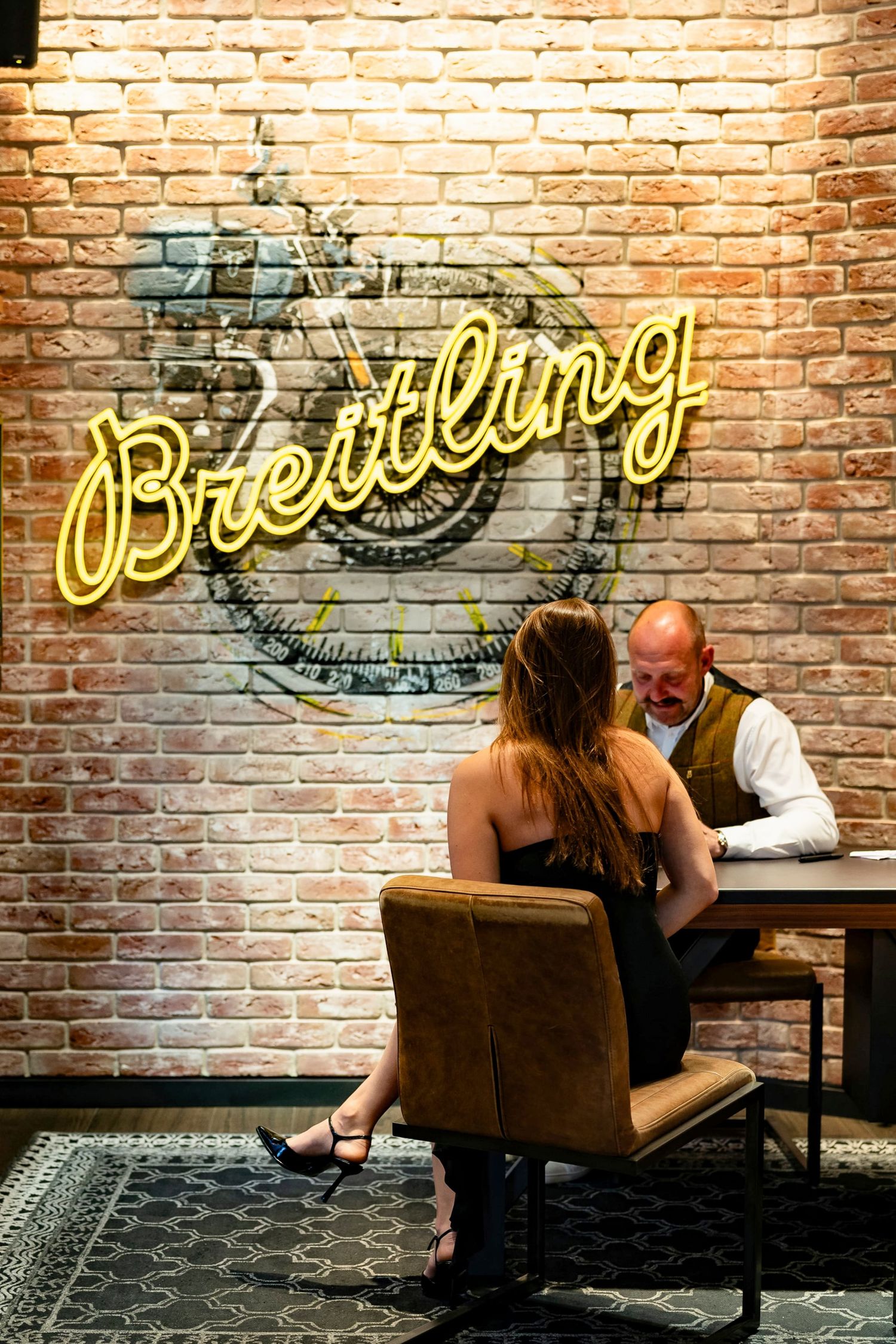 Couple sitting at a table beneath a glowing Breitling neon sign on a rustic brick wall.