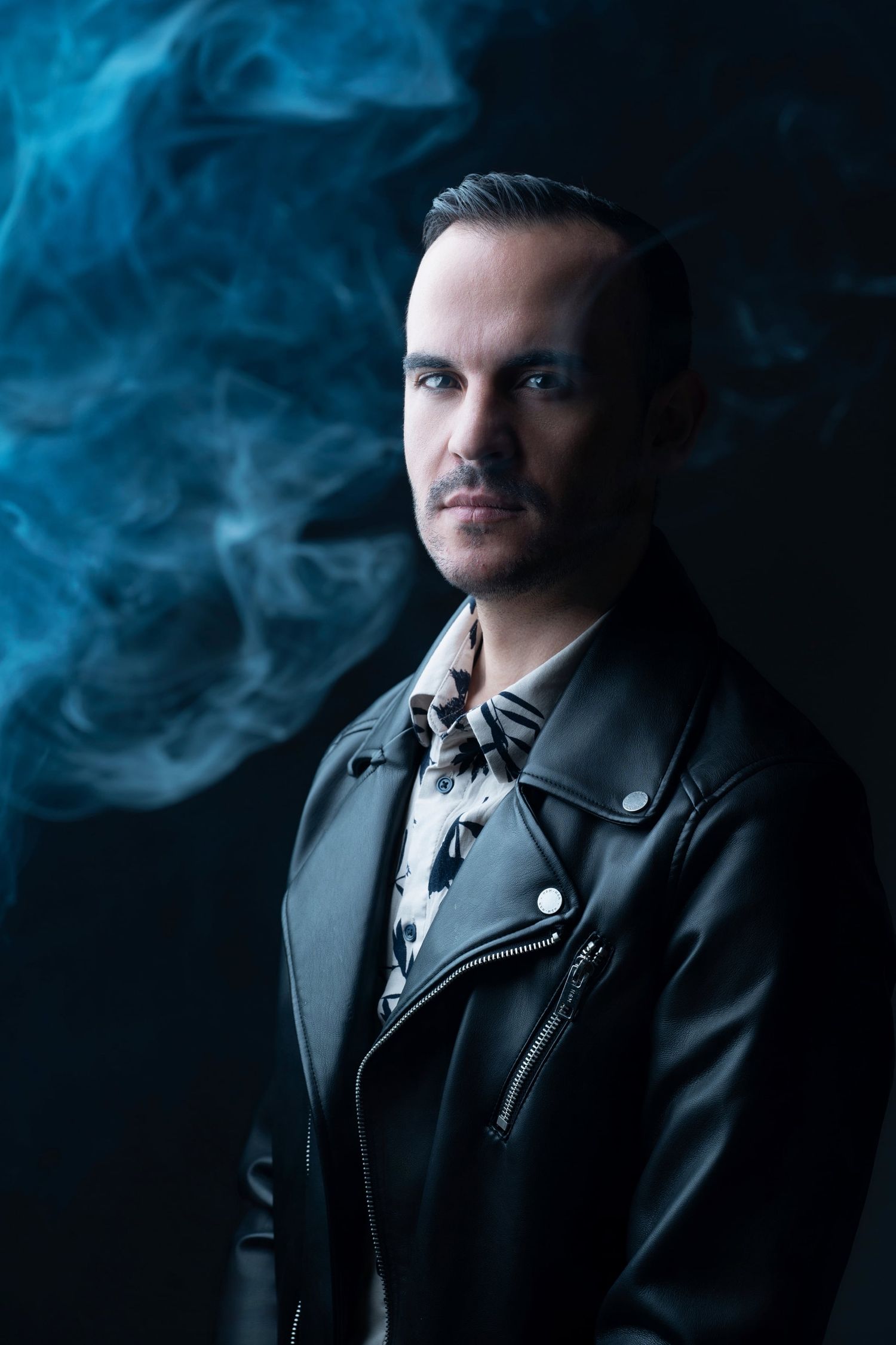 Mysterious man in black leather jacket posing with blue smoke swirling around him on dark background.