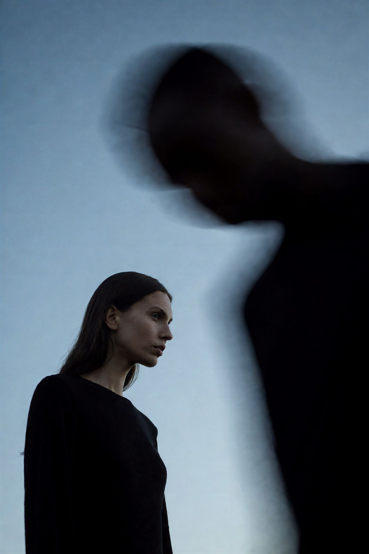 A woman in black stands in focus while a blurred dark figure looms in the foreground.