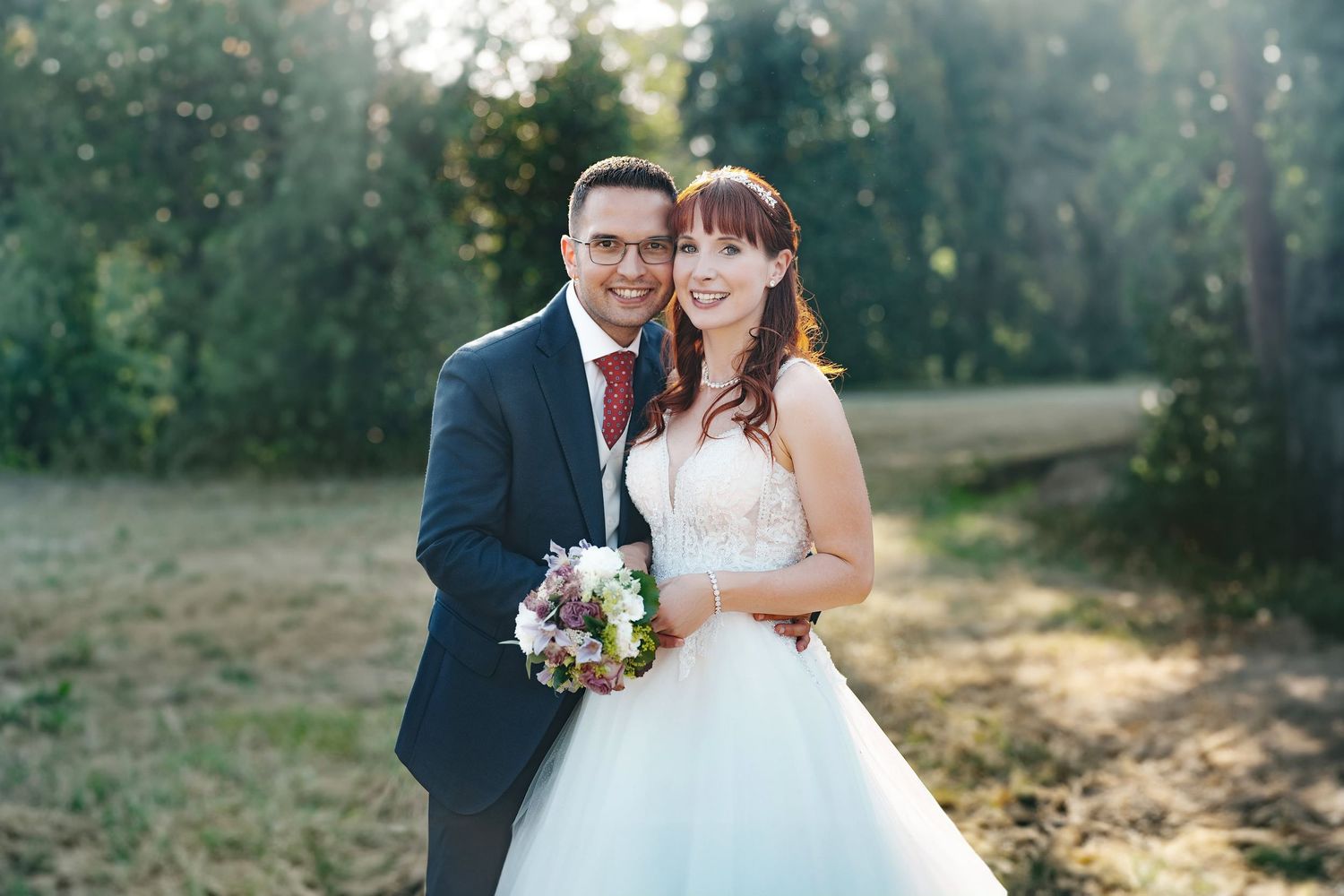 Happy wedding couple posing outdoors in golden sunlight, bride holding a bouquet of flowers.