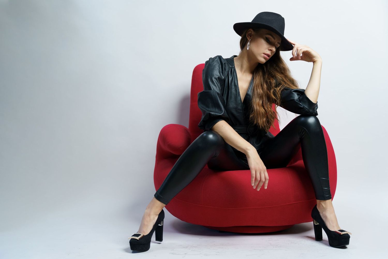 Stylish woman in black leather outfit and wide-brim hat posing on red chair in studio.