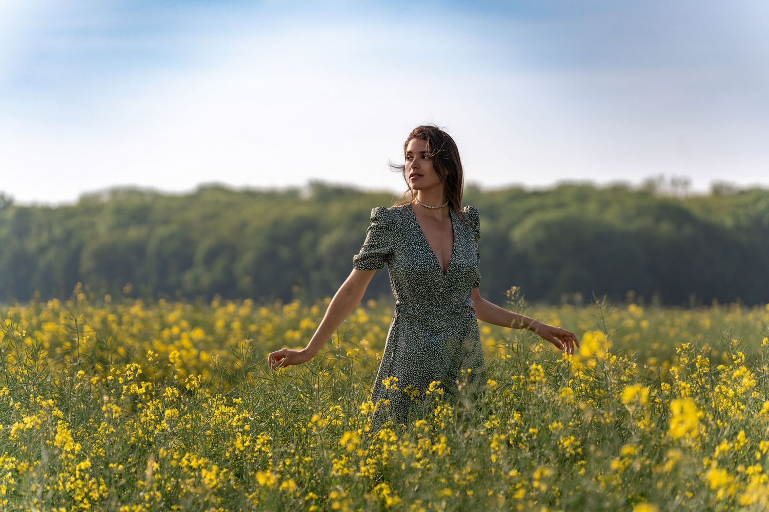 Woman in green floral dress standing in yellow flower field with arms outstretched.