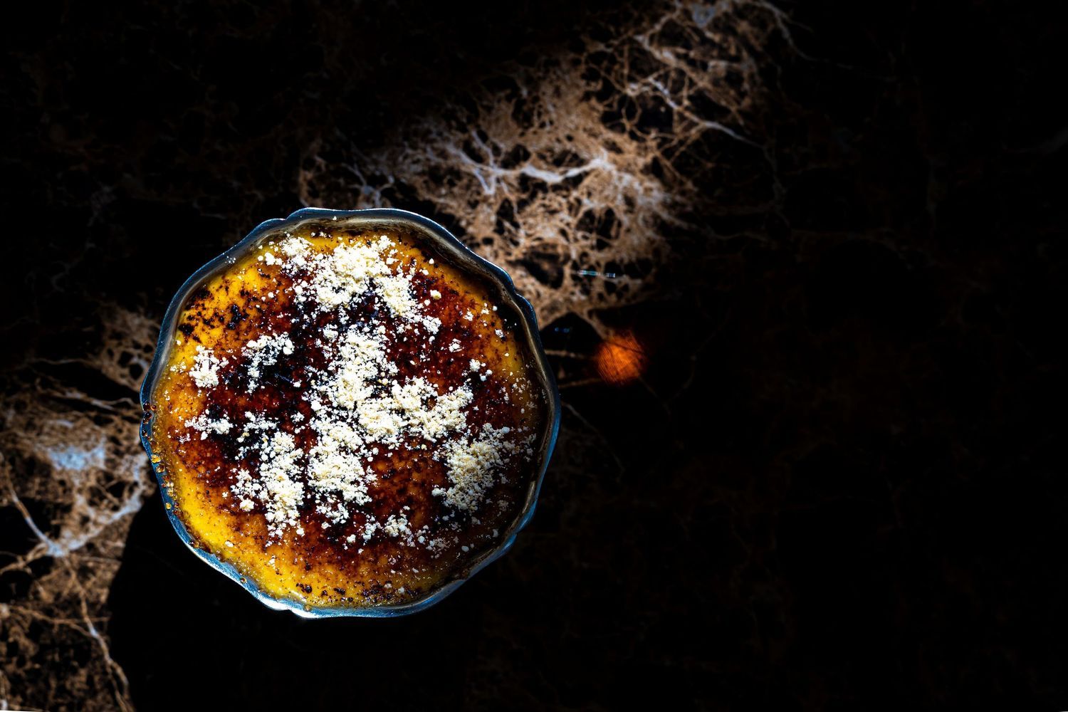 Golden baked dessert topped with powdered sugar in a glass dish on dark marble surface.