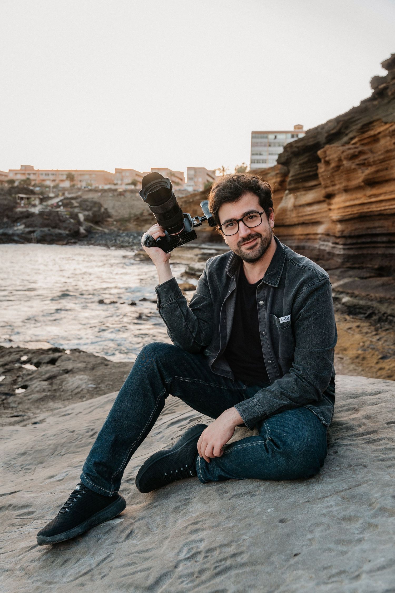 Man with glasses holding camera sitting on coastal rocks outdoors.