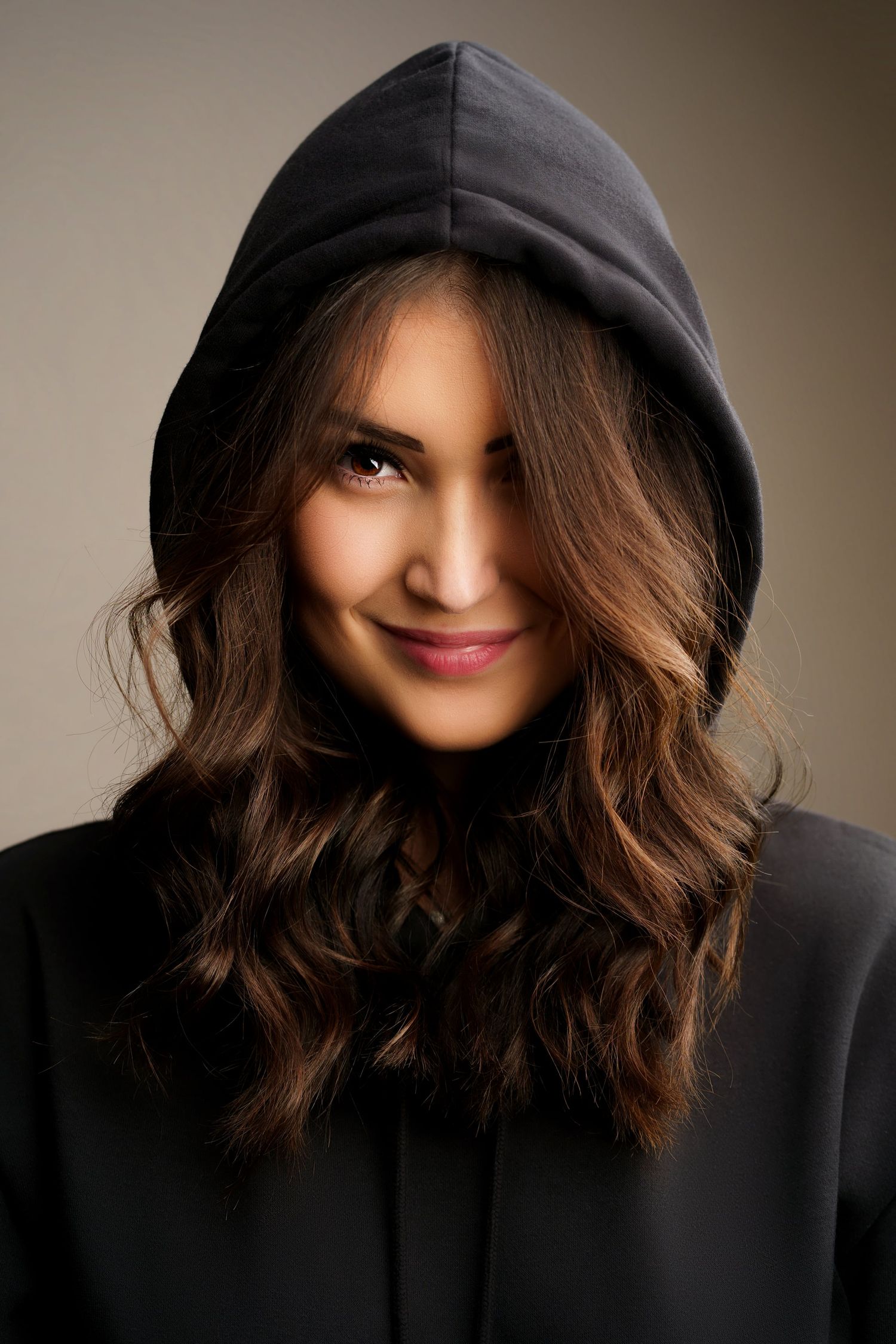 Young woman with wavy brown hair wearing a black hoodie, smiling softly against a neutral background.