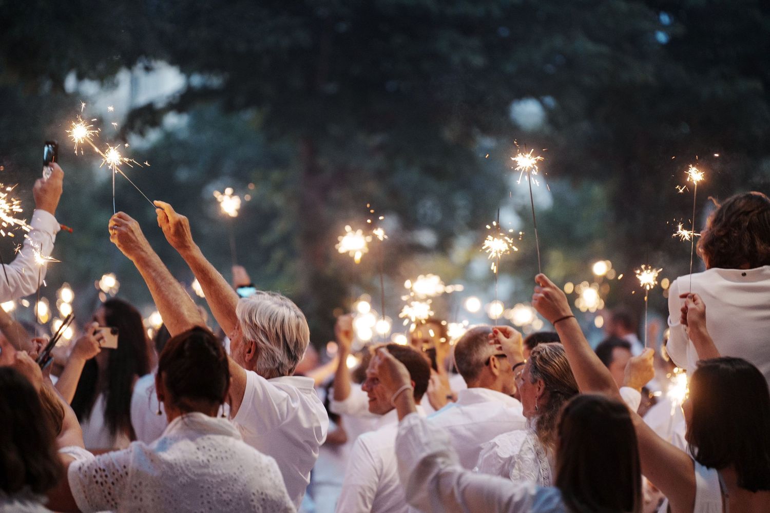 Crowd of people dressed in white celebrating outdoors at night with sparklers raised in the air.